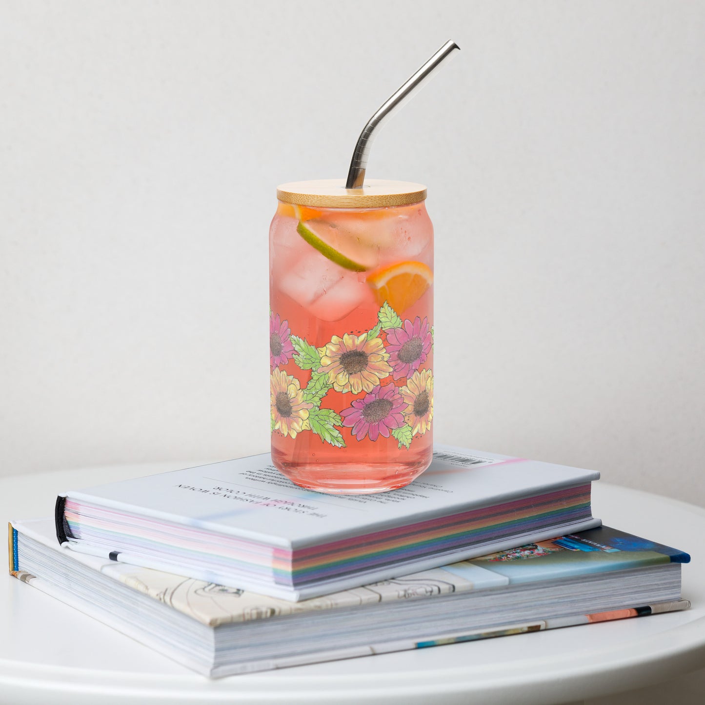 16 ounce can-shaped glass. Features wraparound print of watercolor Gerber daisies. Has a bamboo lid and stainless steel straw.  Shown filled with pink lemonade and sitting on a stack of books.