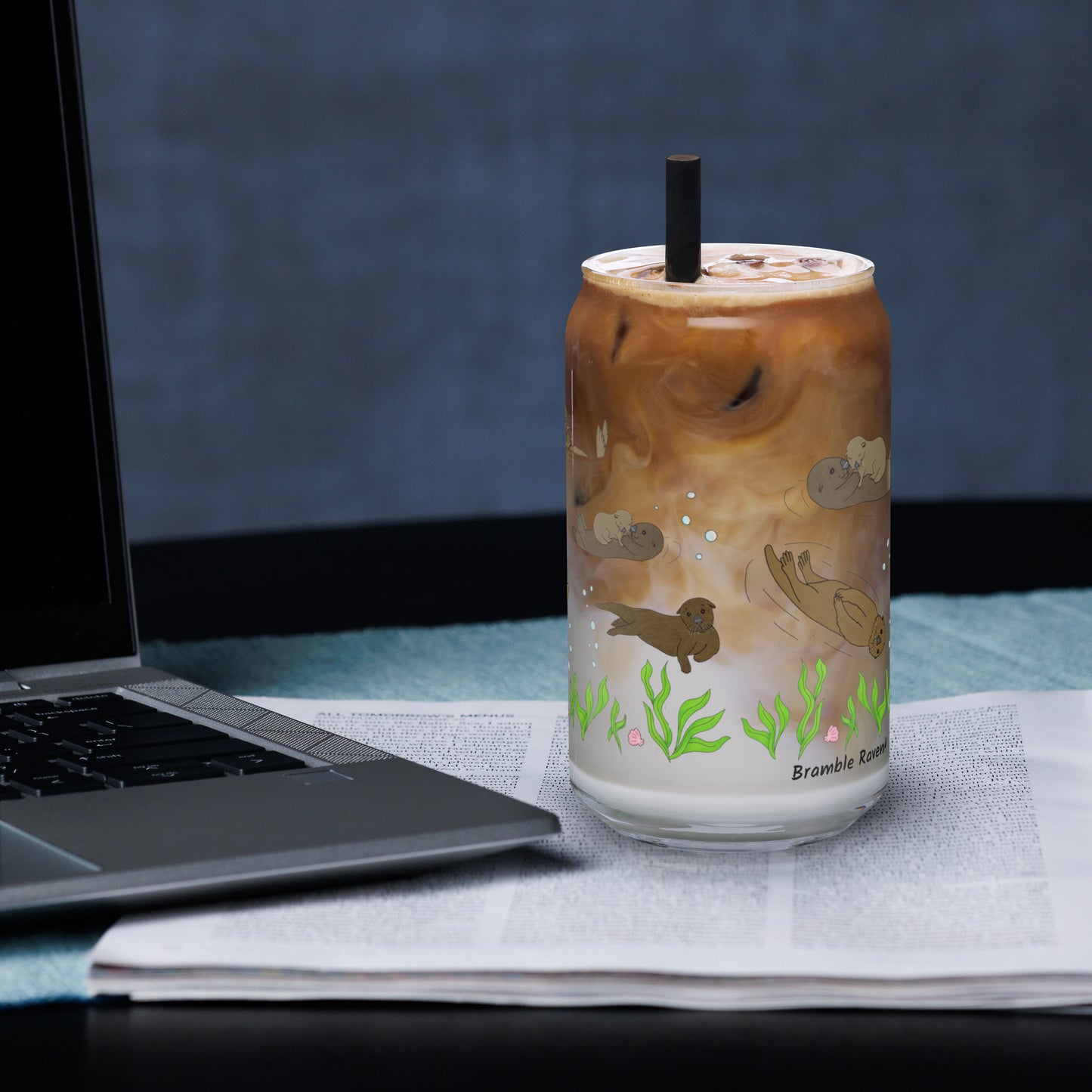 Can-shaped glass holds 16 fluid ounces. Has a design of sea otters swimming above the seaweed with bubble and shell accents. Filled with iced coffee and a straw, sitting by a laptop.