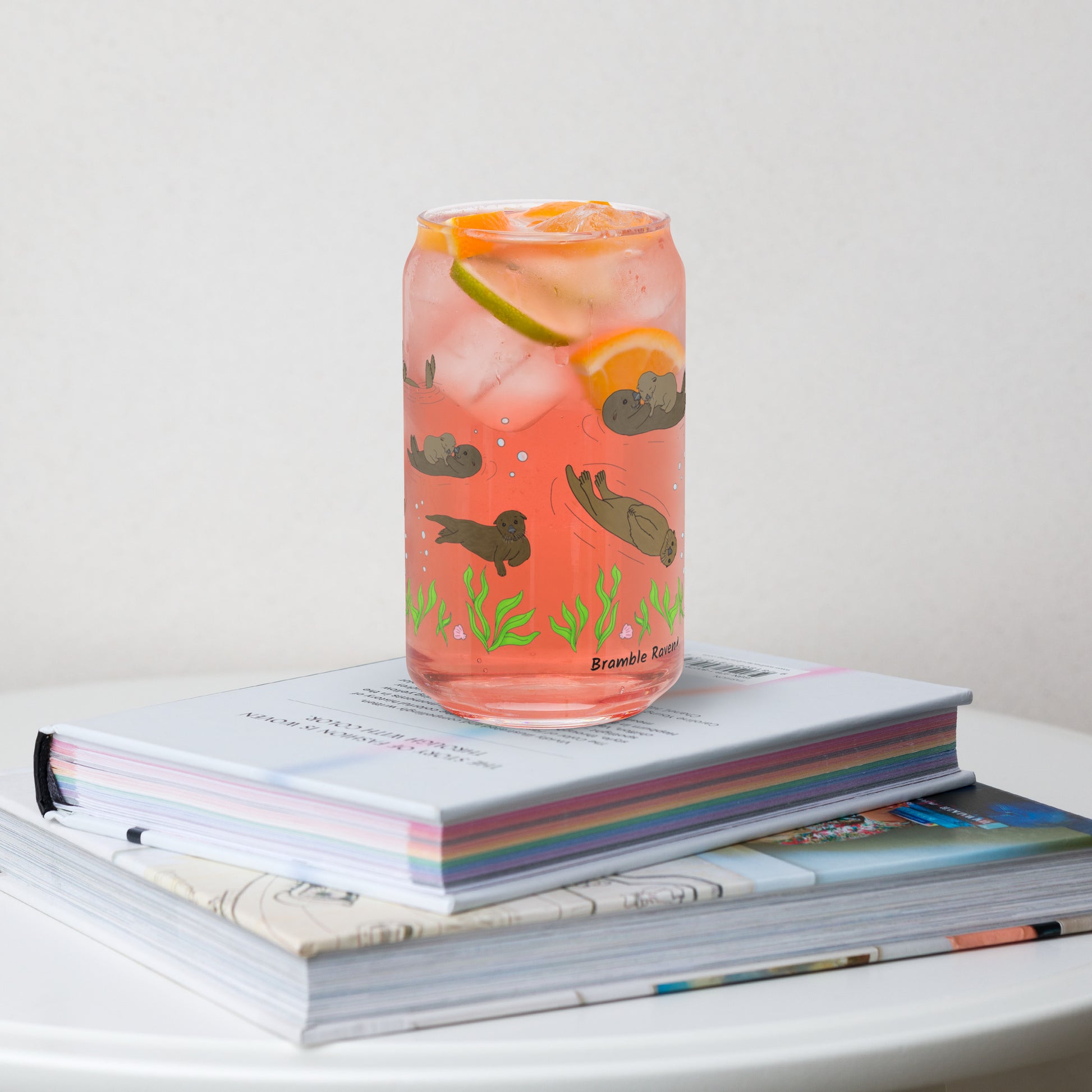 Can-shaped glass holds 16 fluid ounces. Has a design of sea otters swimming above the seaweed with bubble and shell accents. Shown filled with pink lemonade and citrus wedges, sitting on a stack of books.