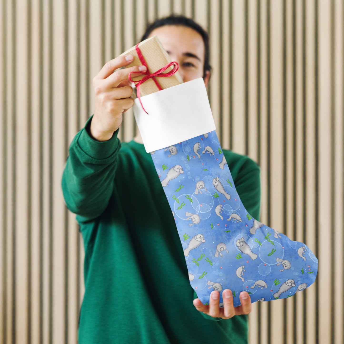Limited Edition Manatee Christmas Stocking.  7 by 18 inches. The front has a hand-illustrated manatee design with an off-white polyester fabric on the back. It has a white fold-over cuff and a loop for hanging. Shown being held by male model.