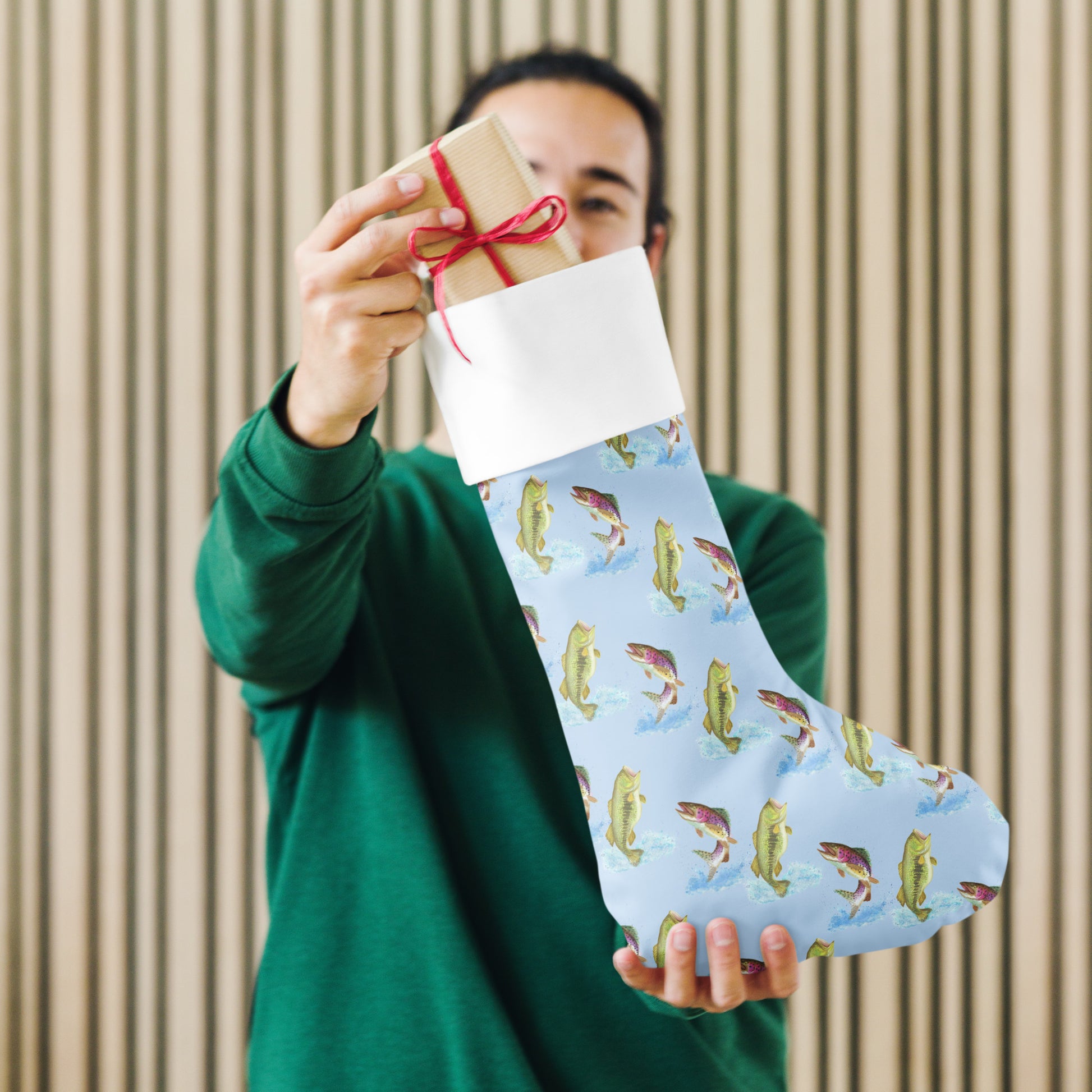 Limited Edition Blue Fishing Christmas Stocking.  7 by 18 inches. The front has watercolor rainbow trout and largemouth bass patterned on a blue background and an off-white polyester back fabric. It has a white fold-over cuff and a hanging loop. Shown being held by male model.