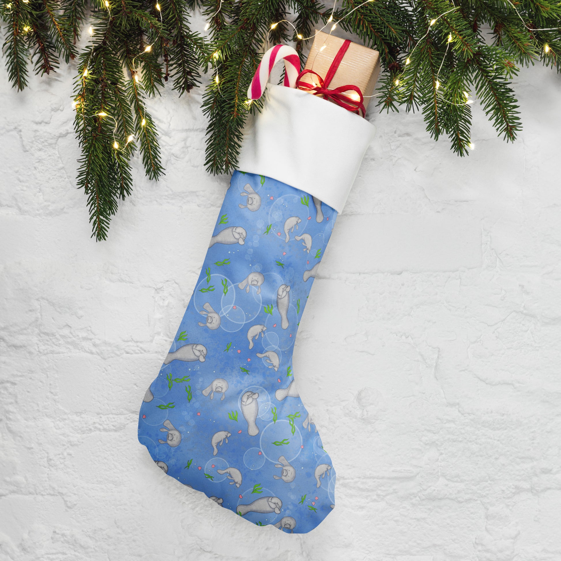 Limited Edition Manatee Christmas Stocking.  7 by 18 inches. The front has a hand-illustrated manatee design with an off-white polyester fabric on the back. It has a white fold-over cuff and a loop for hanging. Shown stuffed with gifts by pine boughs.