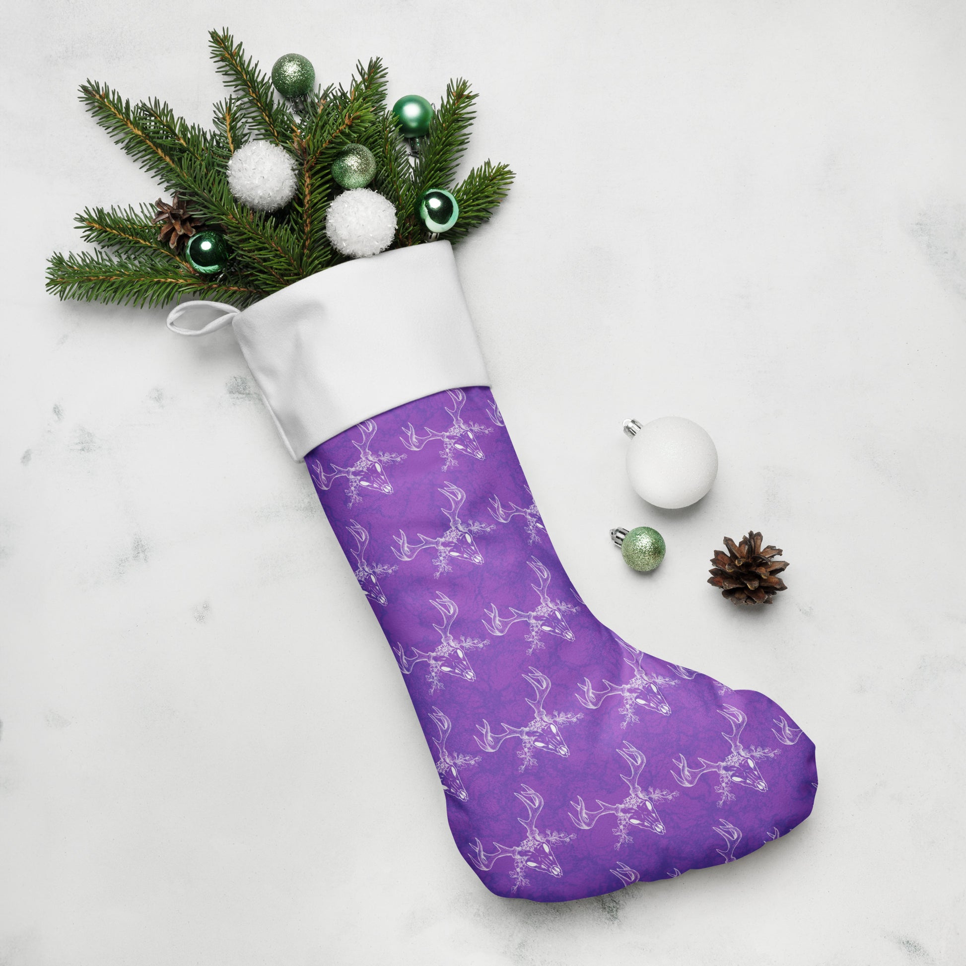 Limited Edition Purple Deer Skull Christmas Stocking.  7 by 18 inches. The front has a hand-illustrated deer skull design on a purple background with an off-white polyester fabric on the back. It has a white fold-over cuff and a loop for hanging. Shown stuffed with pine boughs and ornaments.