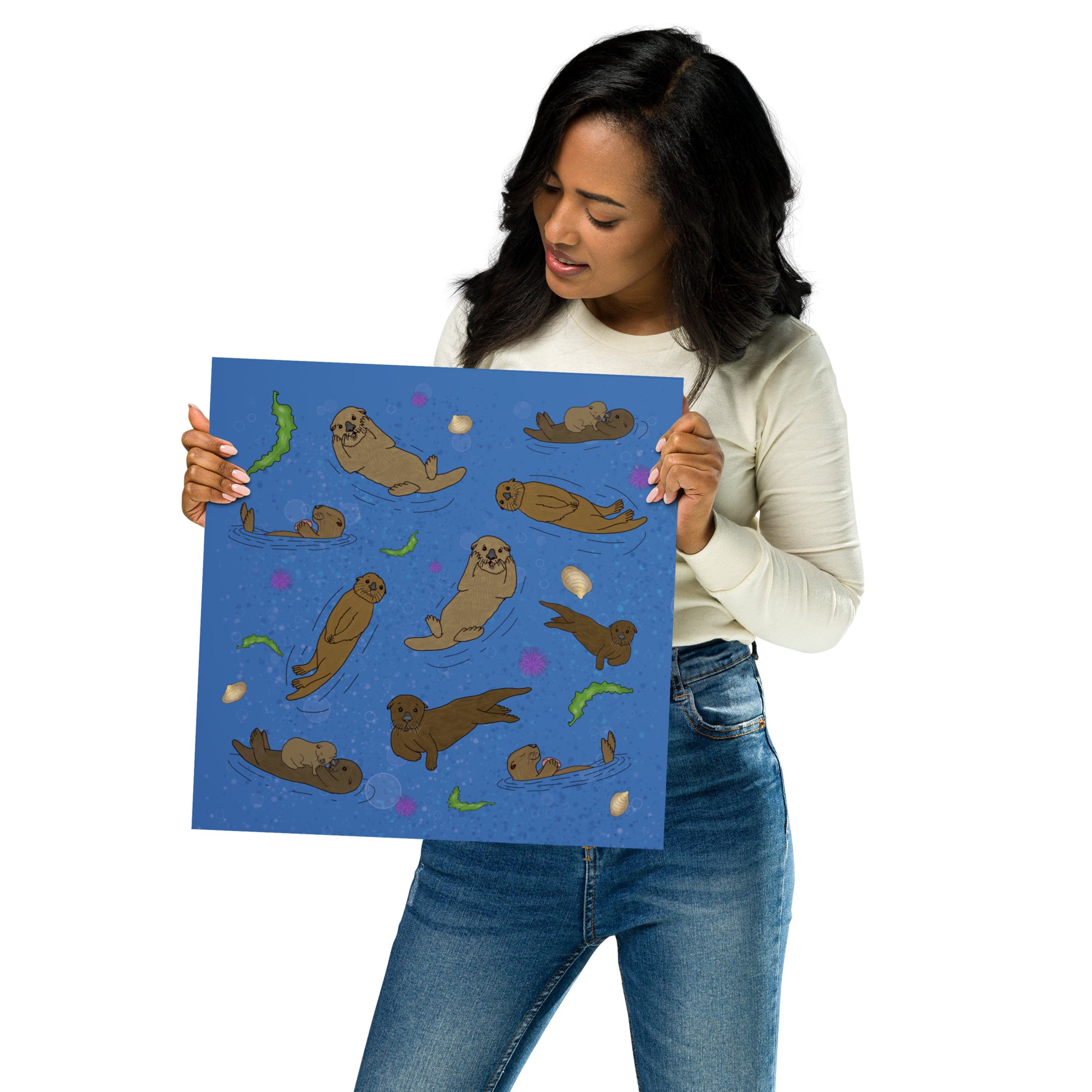 14 x 14 inch matte poster art print. Hand illustrated digital drawing of cute sea otters with accenting seashells, seaweed, and sea urchins on a blue background. Shown in female model's hands.