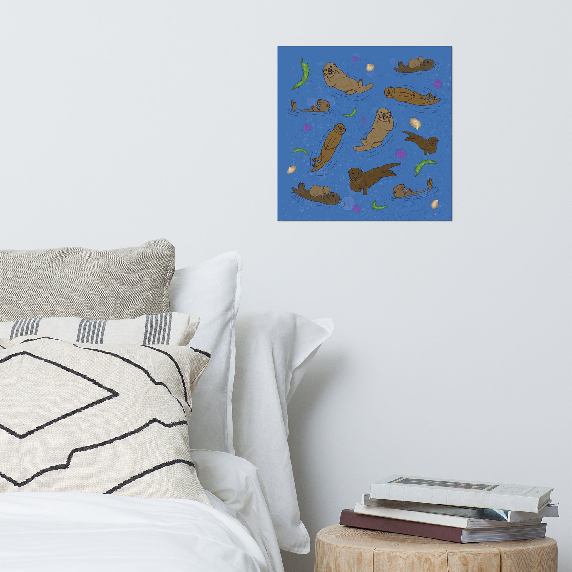 16 x 16 inch matte poster art print. Hand illustrated digital drawing of cute sea otters with accenting seashells, seaweed, and sea urchins on a blue background. Shown on wall above bed and nightstand with books.