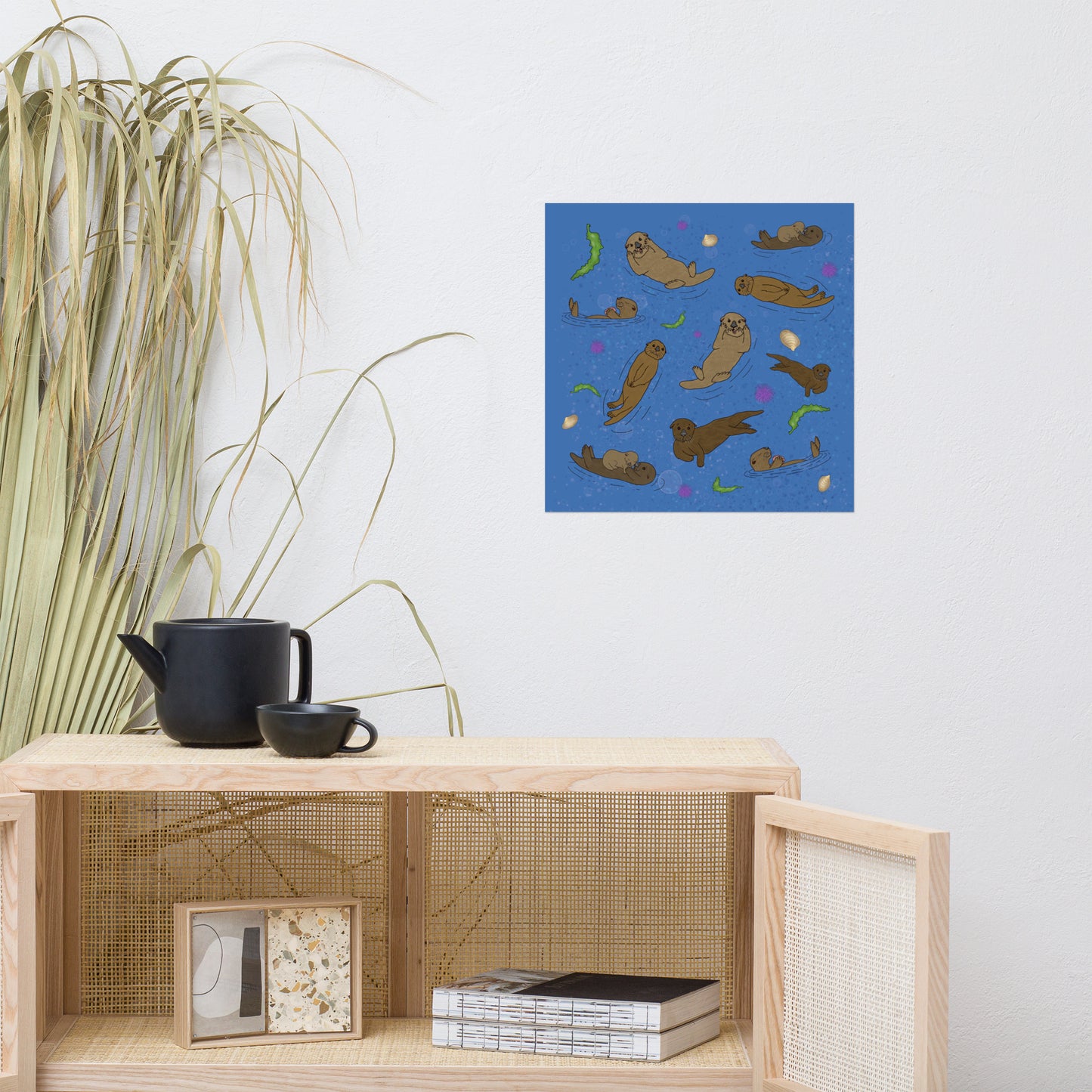 16 x 16 inch matte poster art print. Hand illustrated digital drawing of cute sea otters with accenting seashells, seaweed, and sea urchins on a blue background. Shown on wall above wicker cabinet with black tea pot and grass decor.