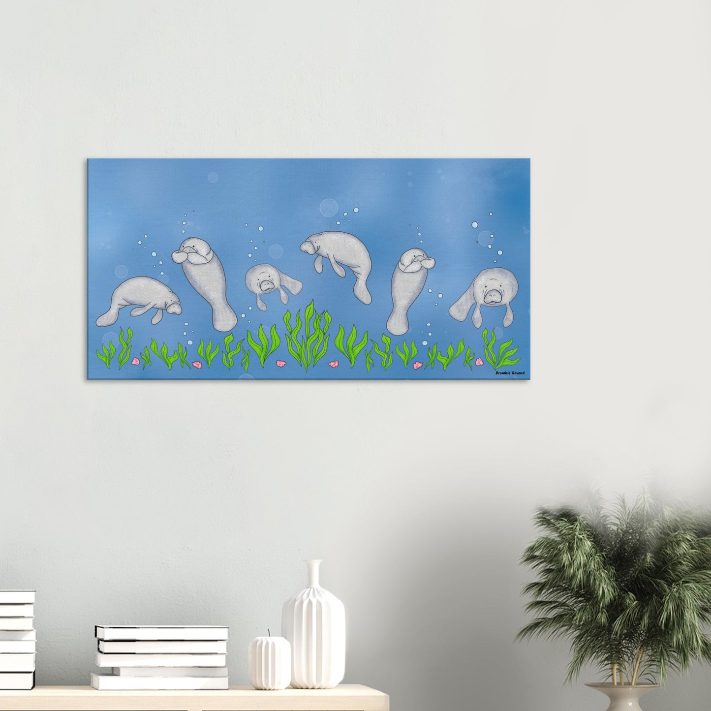 20 by 40 inch slim canvas wall art print featuring cute illustrated manatees swimming above the seabed. Shown on wall above tabletop with books and a potted plant.