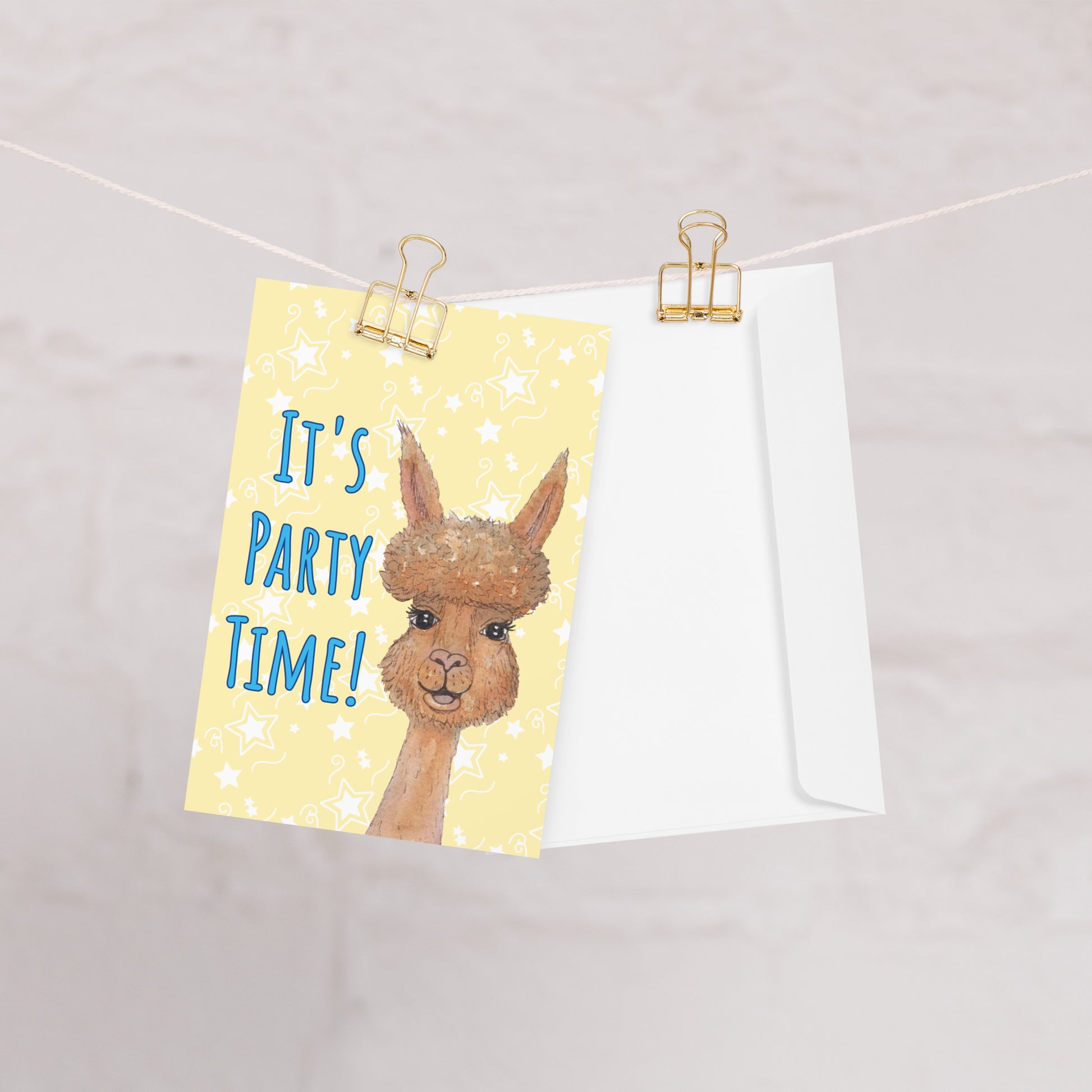 4 by 6 inch party alpaca greeting card and envelope. Image shows card and envelope hanging by clips on string. Background of card is pastel yellow with white star accents. Front has watercolor alpaca and text: It's party time!