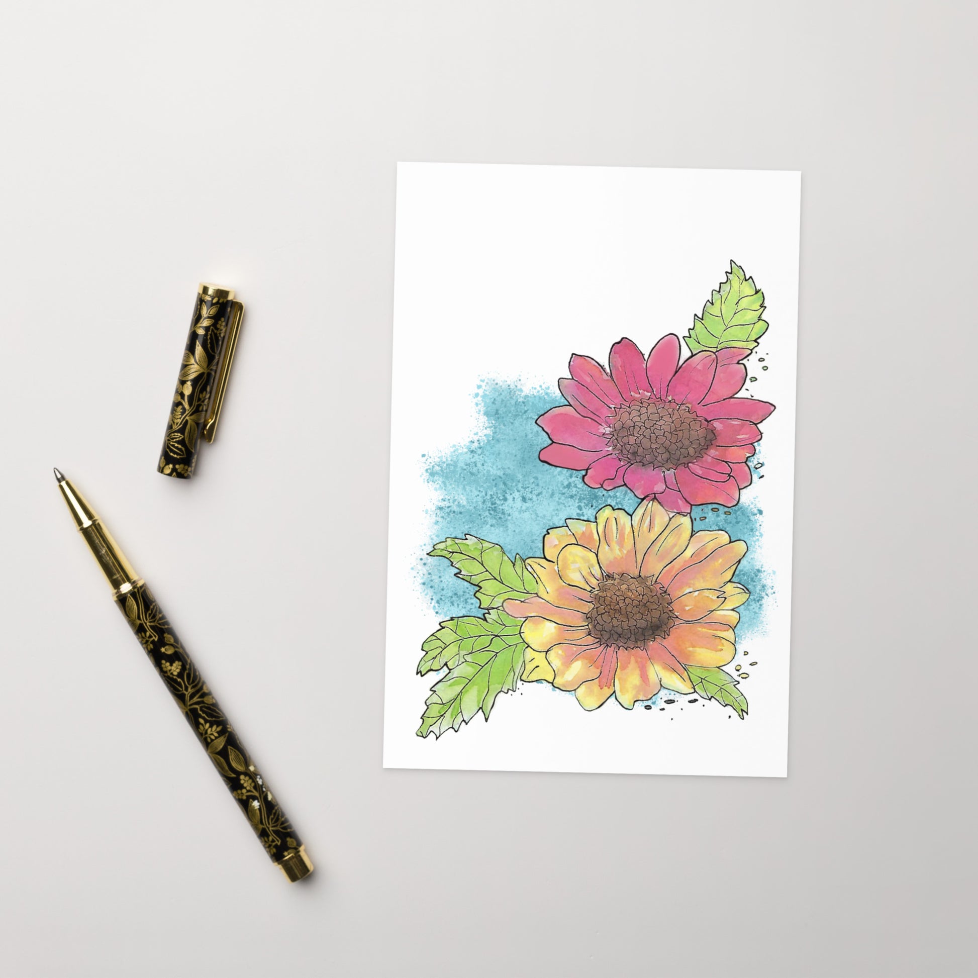 4 by 6 inch watercolor Gerber daisies greeting card and envelope. Has watercolor daisies on the front. The inside has a blue watercolor border with daisy accents and room for a message. Image shows card front by ballpoint pen.