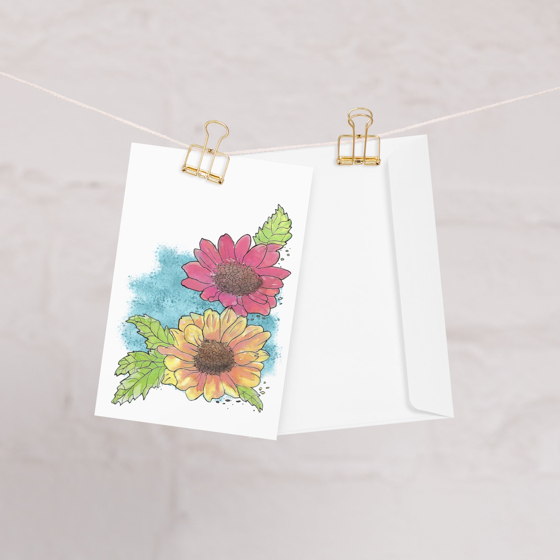 4 by 6 inch watercolor Gerber daisies greeting card and envelope. Has watercolor daisies on the front. The inside has a blue watercolor border with daisy accents and room for a message. Card and envelope shown hanging from clips on a string.