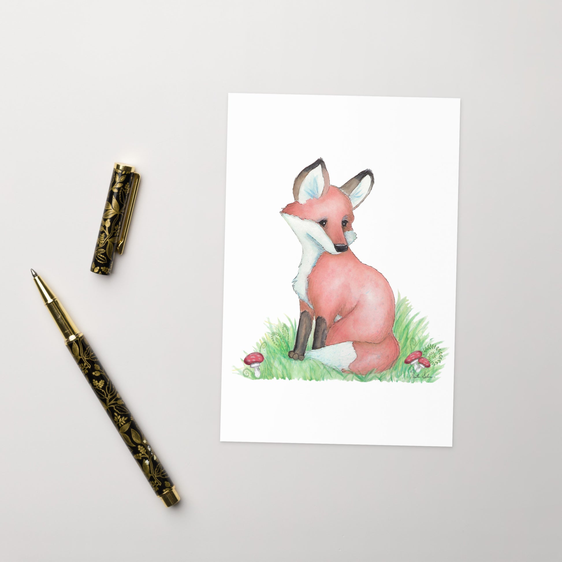 4 by 6 inch forest fox greeting card. Front has watercolor print of a fox in the grass by mushrooms and ferns. Inside is blank. Comes with a white envelope. Made of durable paperboard with vibrant printing. Shown on tabletop by pen.