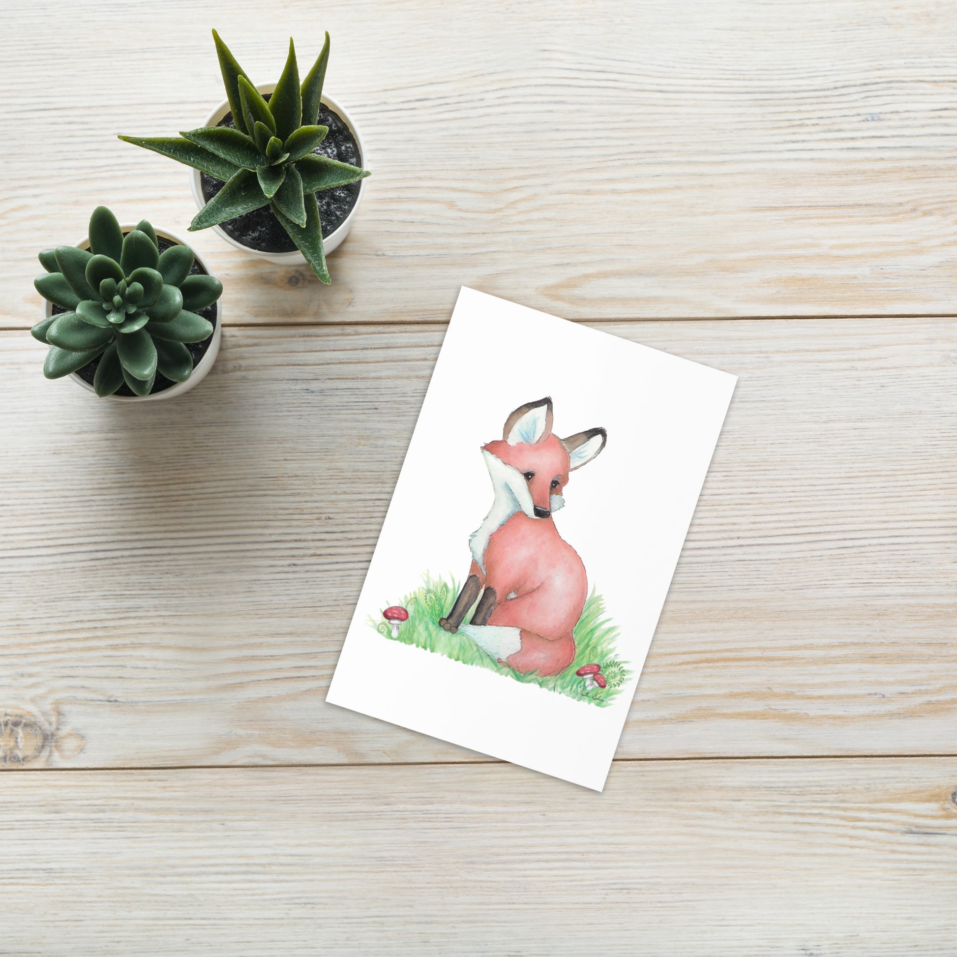 4 by 6 inch forest fox greeting card. Front has watercolor print of a fox in the grass by mushrooms and ferns. Inside is blank. Comes with a white envelope. Made of durable paperboard with vibrant printing. Shown on tabletop by succulents.