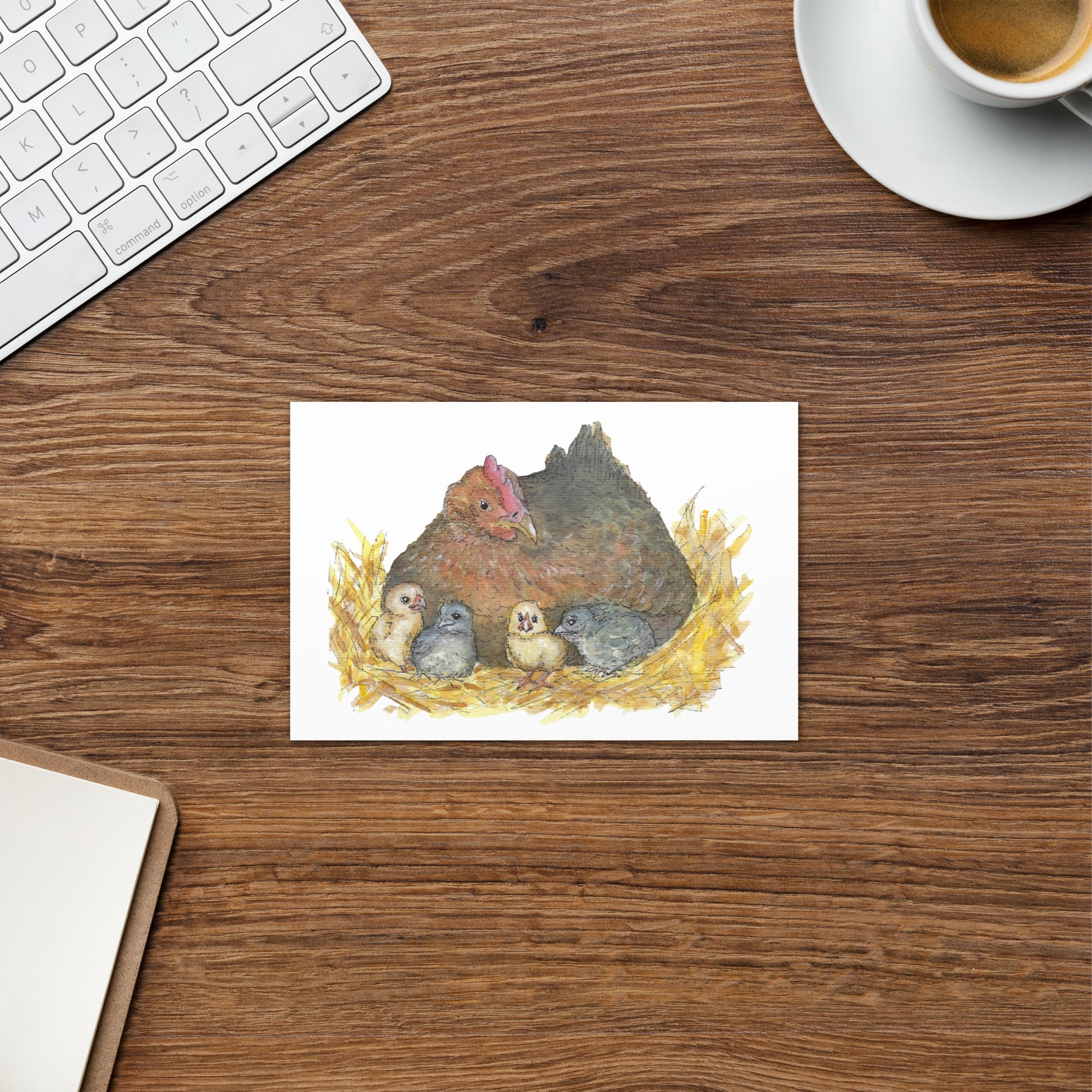 4 by 6 inch greeting card and envelope. Front features watercolor print of a mother hen and four chicks in a nest. Inside is blank. Shown on wooden table by coffee cup and keyboard.