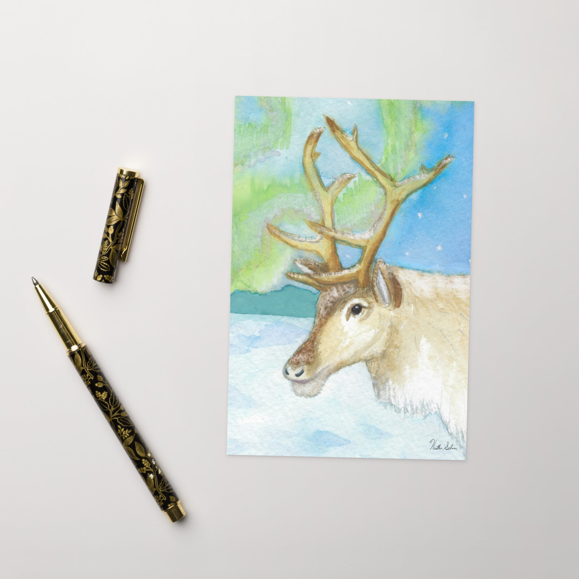 4 x 6 inch greeting card. Features watercolor print of a reindeer in the snow with northern lights in the sky. Blank inside. Comes with a white envelope. Shows front and back cover. Shown on table by pen.