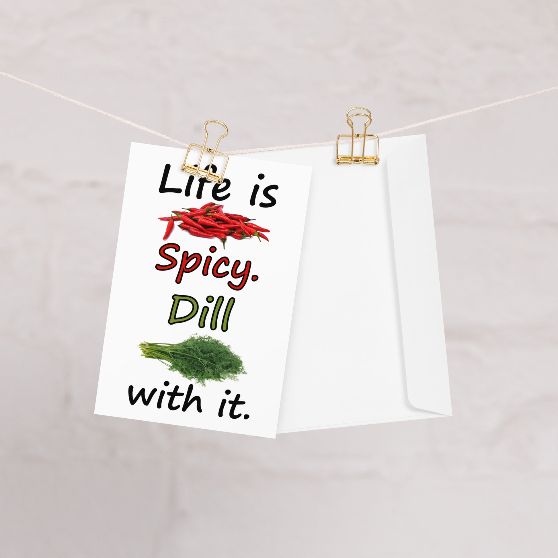 4 by 6 inch Life Is Spicy greeting card. Features Life is Spicy, Dill With It text, with image of chili peppers and dill weed on the front. The inside is blank. Comes with a white envelope. Card is made of durable paperboard with vibrant printing. Card shown on clothesline with white envelope.