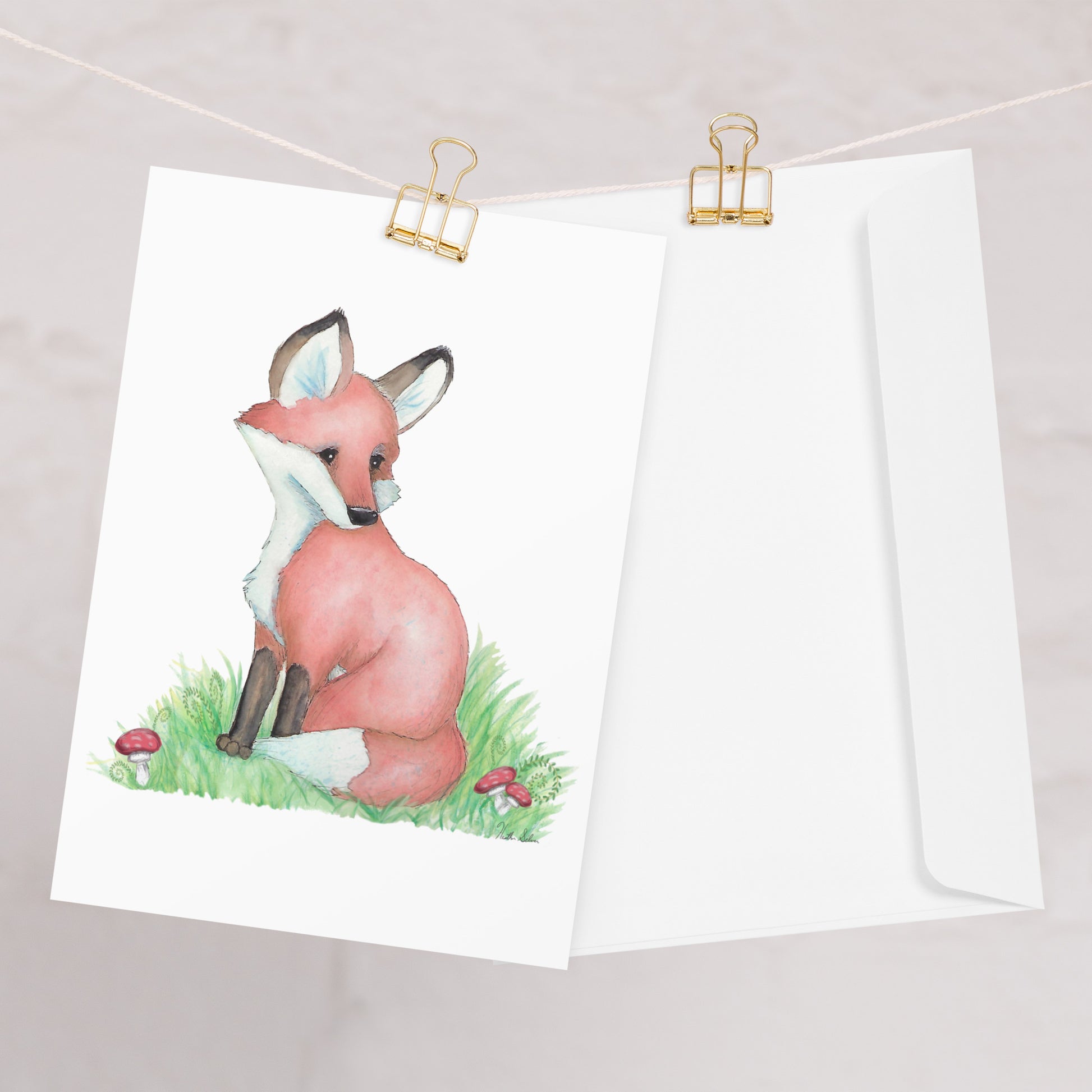Pack of ten 5.5 x 8.5 inch greeting cards with envelopes. Front of card has watercolor fox nestled in the grass by mushrooms and ferns. Inside is blank. Cards are 130 lb 350 gsm coated paperboard with vibrant printing.  One card shown on clothesline with a white envelope.