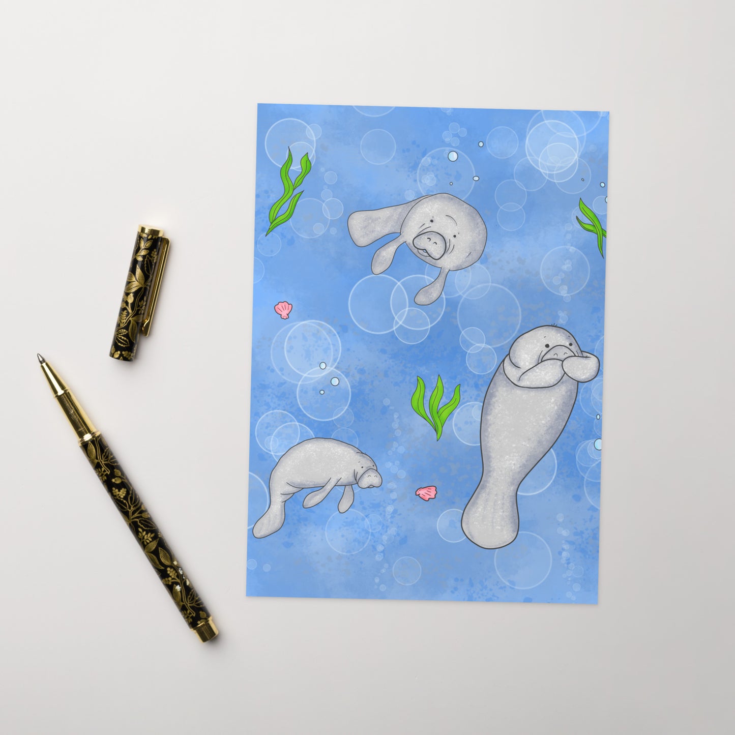Pack of ten 5 x 7 inch greeting cards. Features illustrated roly-poly manatees on the front. Inside is blank. Made of coated paperboard. Comes with ten envelopes. Shown on tabletop by ballpoint pen.