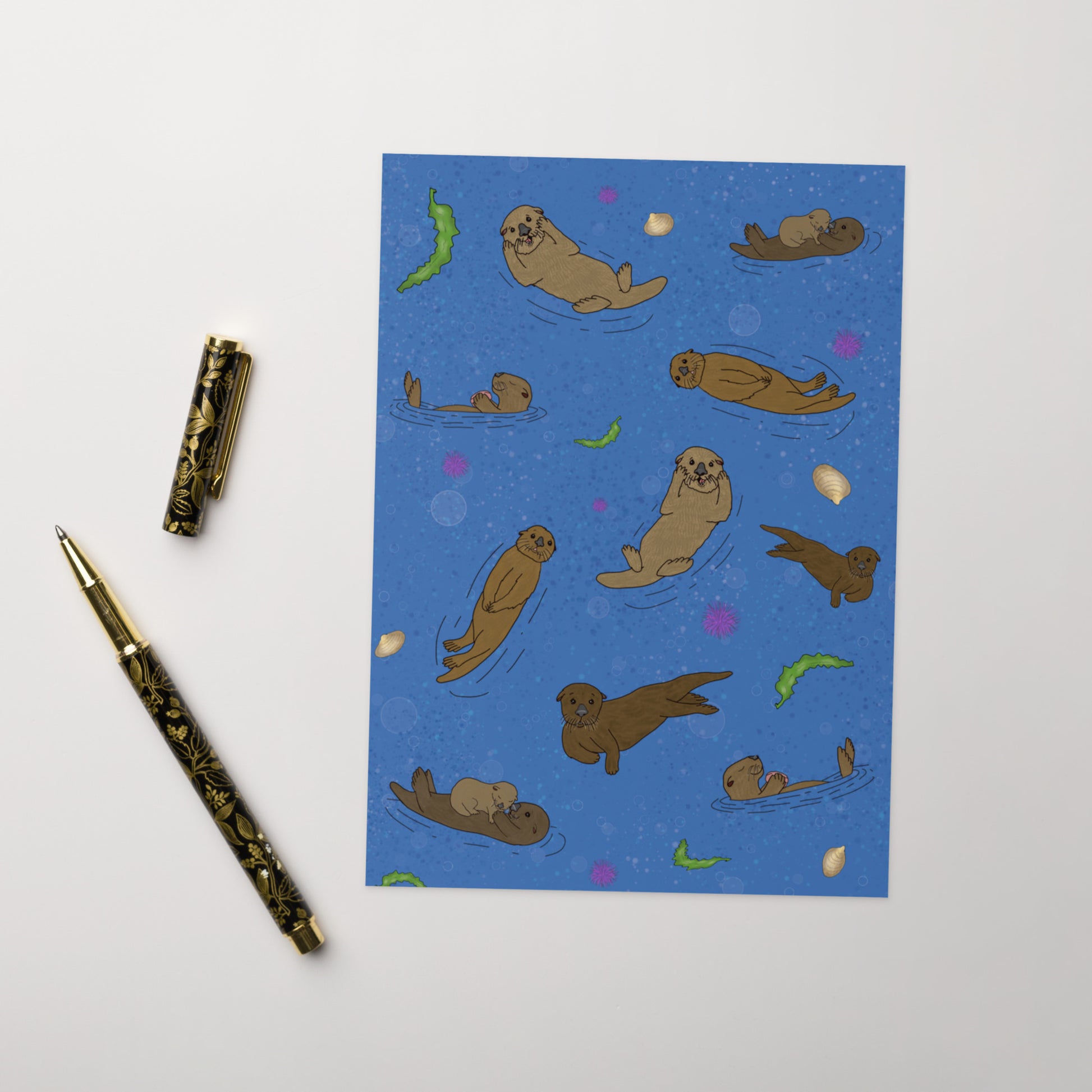 Pack of ten 5 x 7 inch greeting cards. Features illustrated sea otters on the front. Inside is blank. Made of coated paperboard. Comes with ten envelopes. Shown on tabletop by ballpoint pen.