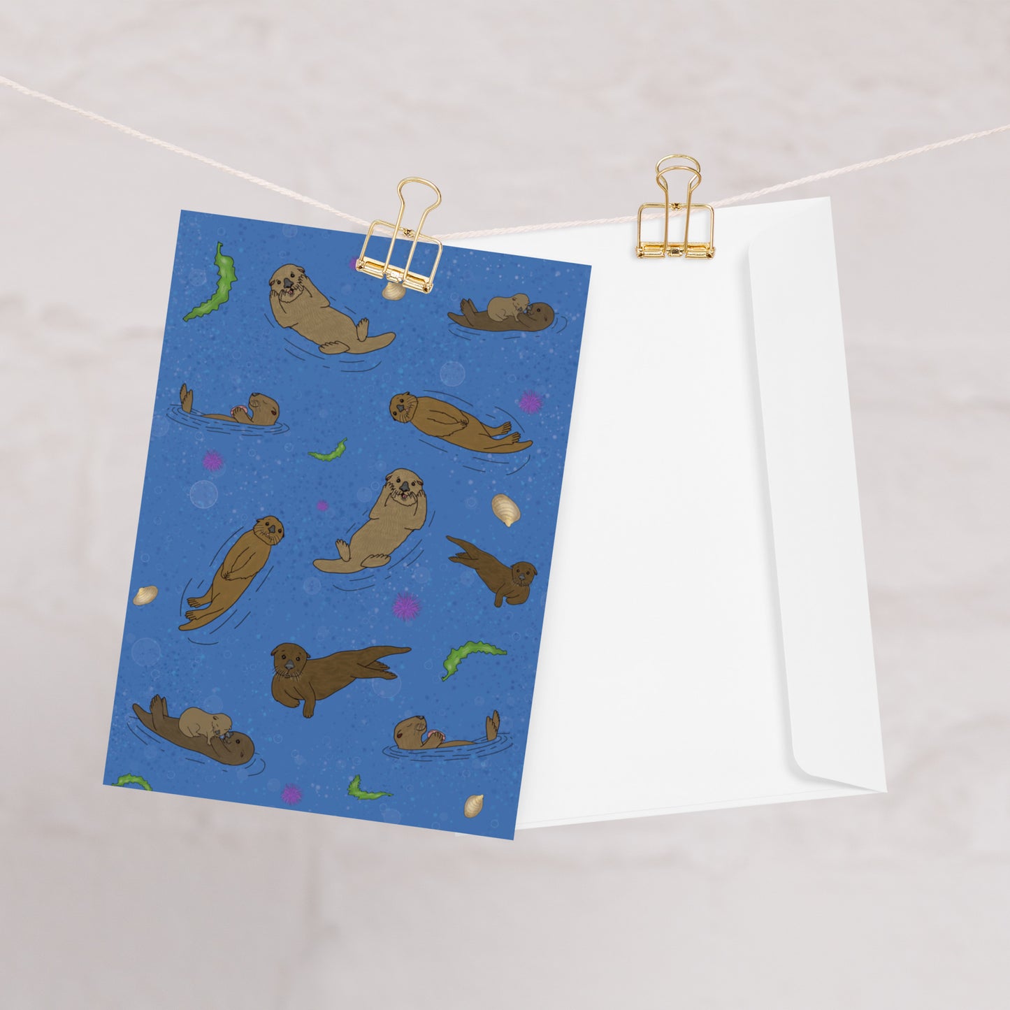 Pack of ten 5 x 7 inch greeting cards. Features illustrated sea otters on the front. Inside is blank. Made of coated paperboard. Comes with ten envelopes. One card shown on clothesline with white envelope.