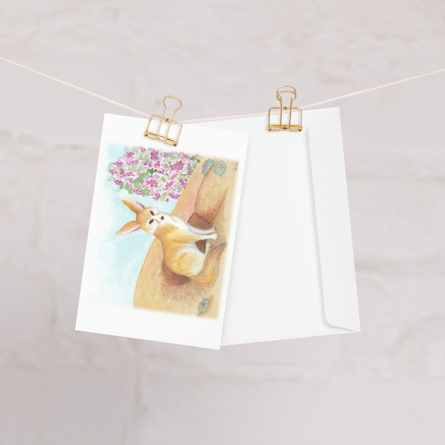 Pack of ten 4 x 6 inch greeting cards. Features watercolor print of fennec fox near it's den in the desert by a jacaranda tree on the front. Inside is blank. Made of coated paperboard. Comes with ten envelopes. One card shown on clothesline with a white envelope.