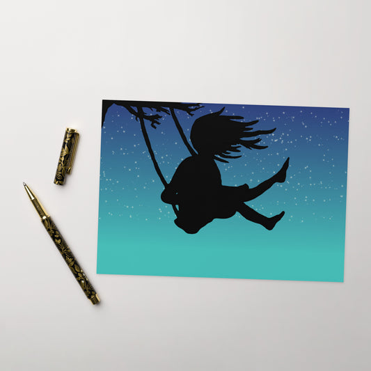 Pack of ten 5.5 by 8.5 inch Night Swing greeting cards and ten envelopes. Front shows silhouette of a girl in a tree swing against a starry summer night sky. Inside is blank. Shown on tabletop by ballpoint pen.