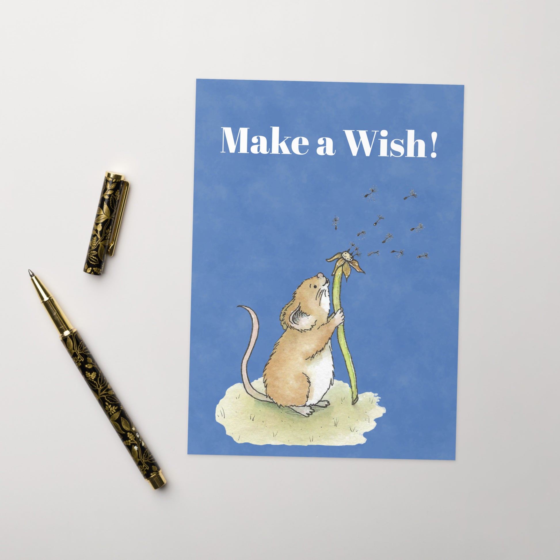 Ten fun greeting cards with ten envelopes. The front features white "Make a wish!" text and our watercolor dandelion wish mouse on a blue background. Inside is blank. Made with thick 350 gsm 150lb coated paperboard with vibrant printing.  Cards measure 5 by 7 inches. Shown on tabletop by ballpoint pen.