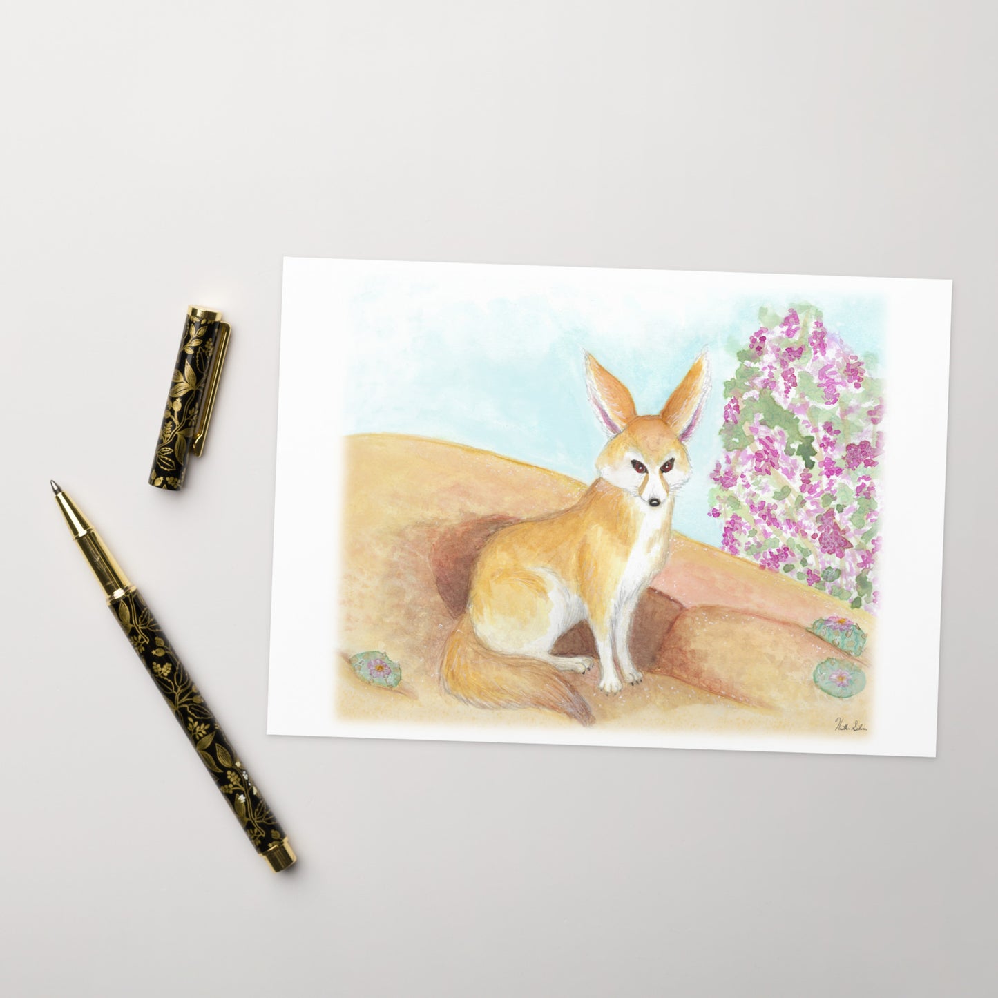 Pack of ten 5 x 7 inch greeting cards. Features watercolor print of fennec fox near it's den in the desert by a jacaranda tree on the front. Inside is blank. Made of coated paperboard. Comes with ten envelopes. Shown on tabletop by ballpoint pen.