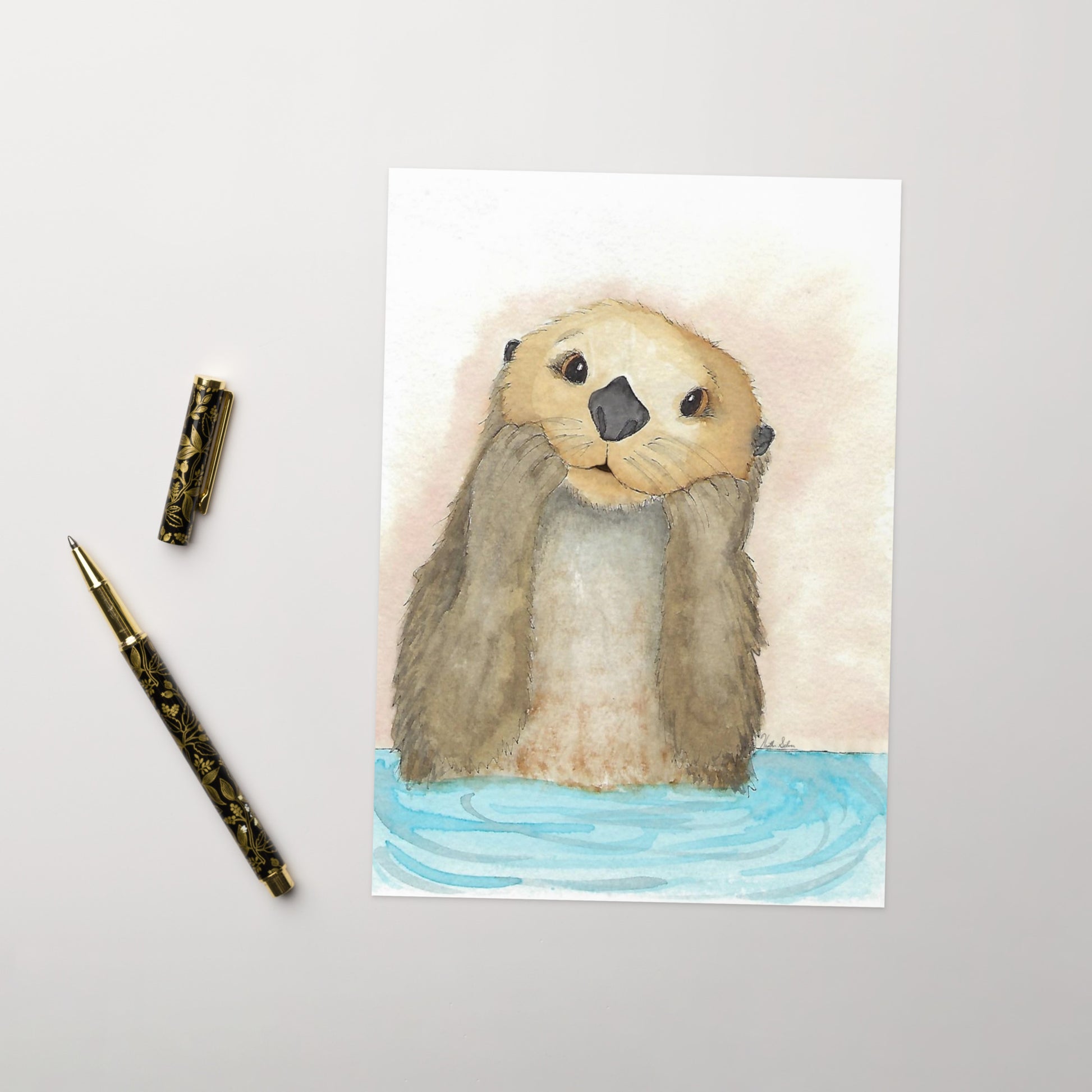 Pack of ten 5 x 7 inch greeting cards. Features watercolor print of a playful sea otter on the front. Inside is blank. Made of coated paperboard. Comes with ten envelopes. Shown on tabletop by ballpoint pen.