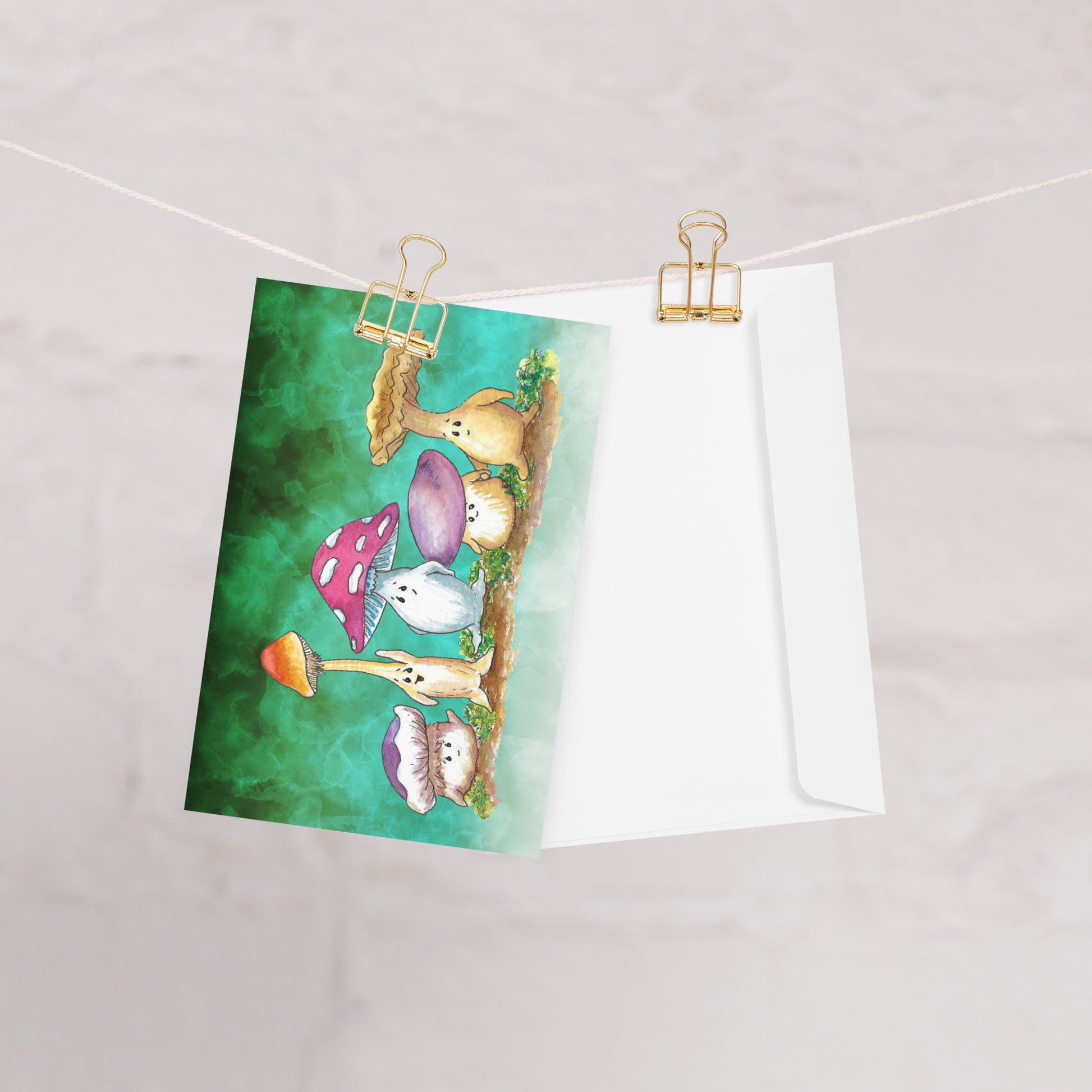 A pack of ten greeting cards and ten envelopes.  Each card is 5  by 7 inches. The front features a watercolor print of Mushy and his whimsical mushroom friends on a green gradient background. The inside has a slim green border and plenty of room for your message. One card shown on a clothesline with a white envelope.