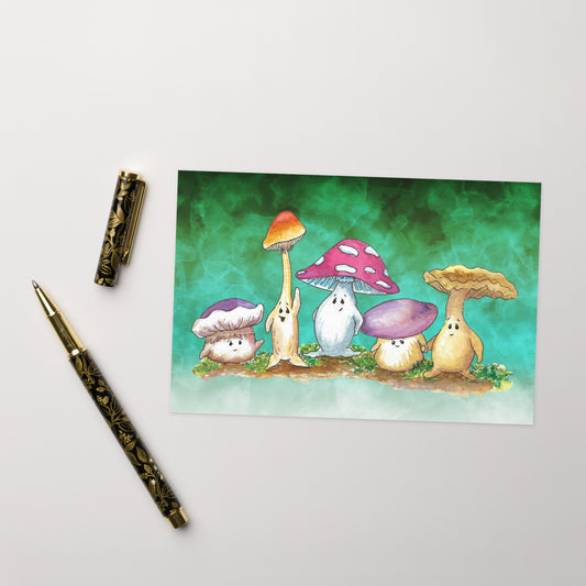 A pack of ten greeting cards and ten envelopes.  Each card is 5  by 7 inches. The front features a watercolor print of Mushy and his whimsical mushroom friends on a green gradient background. The inside has a slim green border and plenty of room for your message. Shown on tabletop by ballpoint pen.