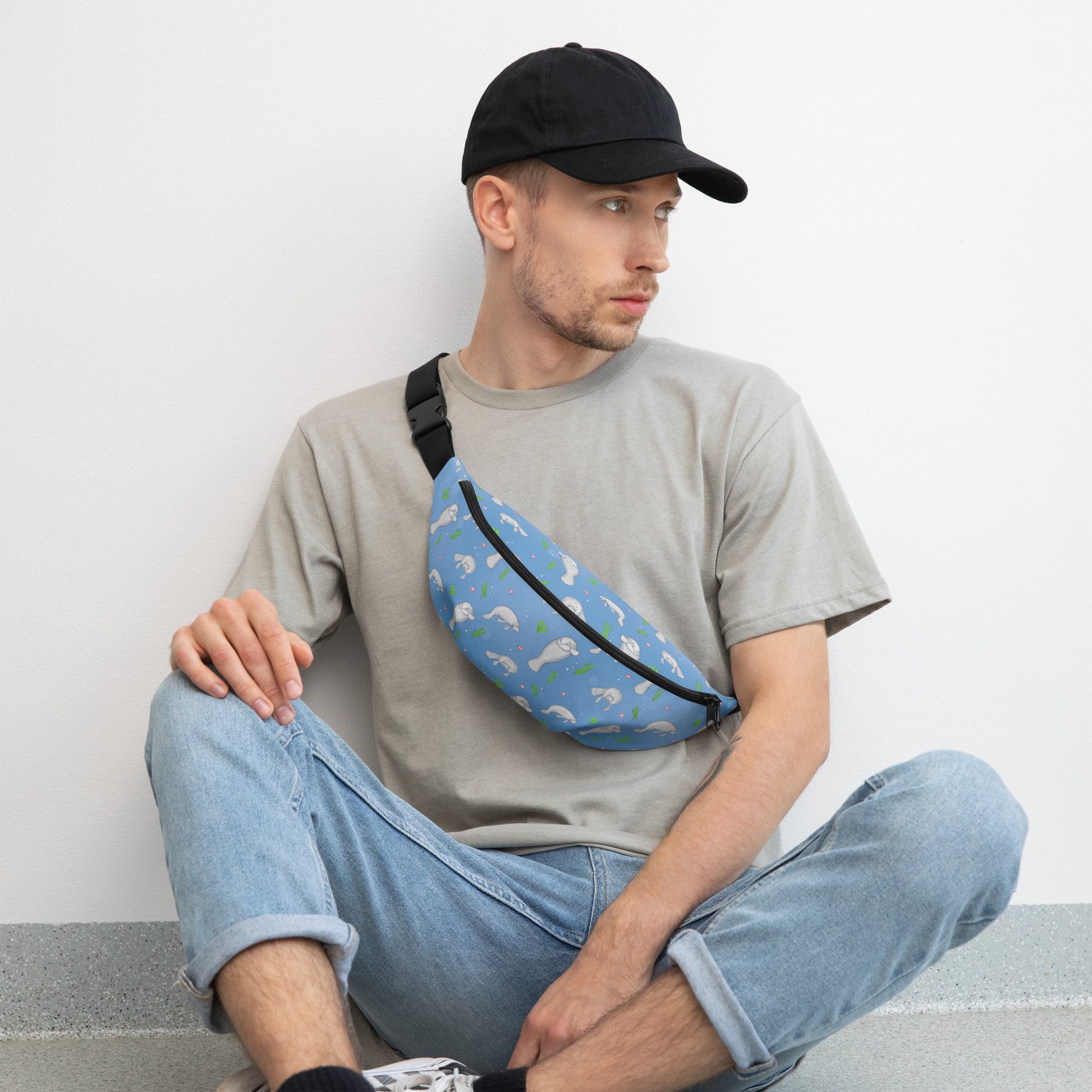 Cute manatee patterned belt bag with adjustable straps, zippered pouch, and small inner pocket. Male model wearing bag across his chest while sitting on the floor.