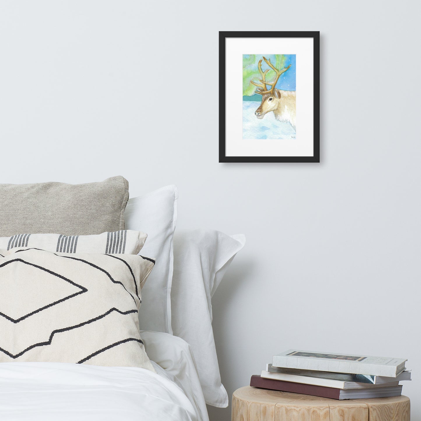 30 by 40 cm art print of a watercolor reindeer in the snow against the northern lights. Framed by a white mat and black ayous wood frame. Has acrylite cover and hanging hardware. Digitally signed by the artist, Heather Silver. Shown on wall above wooden nightstand, books, and bed.