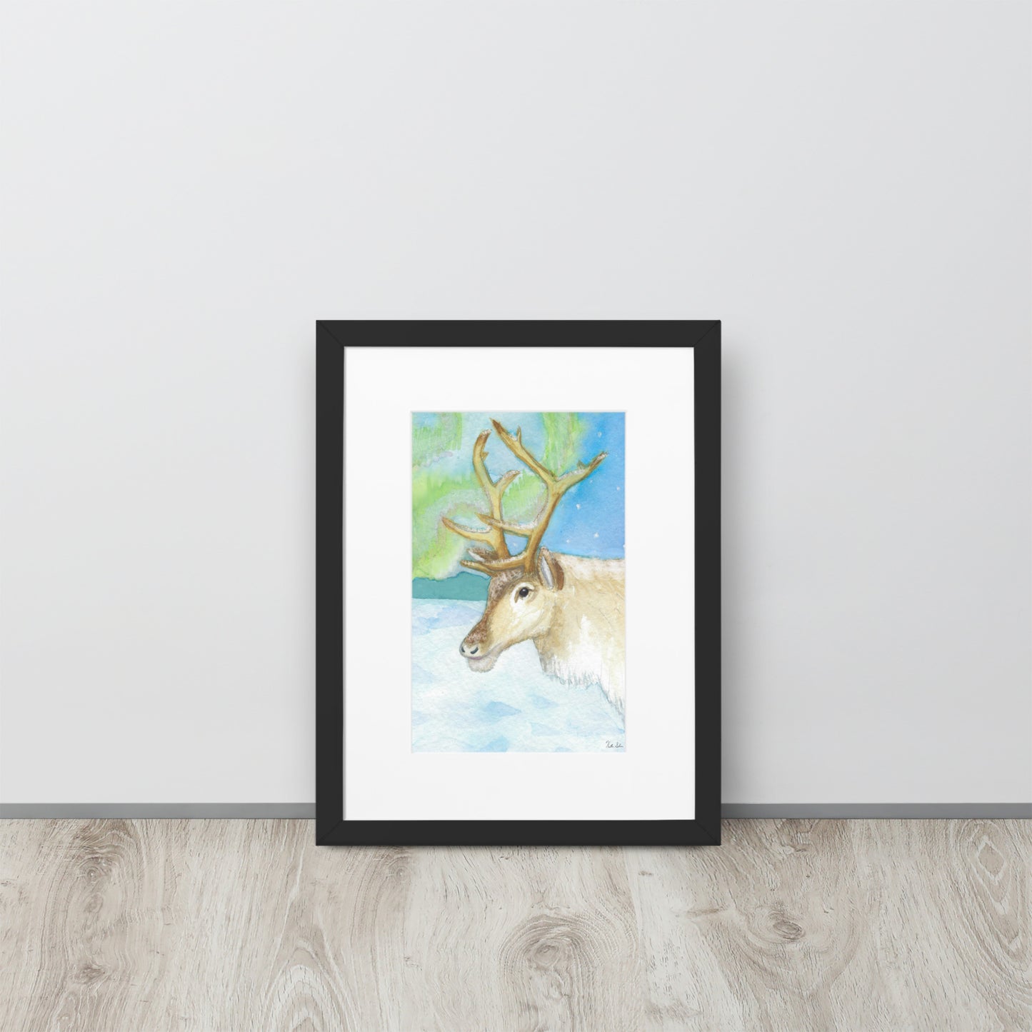 30 by 40 cm art print of a watercolor reindeer in the snow against the northern lights. Framed by a white mat and black ayous wood frame. Has acrylite cover and hanging hardware. Digitally signed by the artist, Heather Silver. Shown leaning against wall on wooden floor.