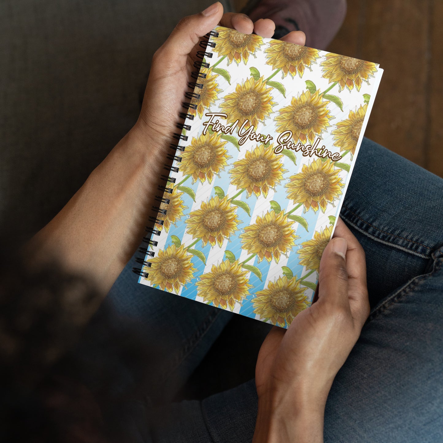 Spiral bound notebook with soft touch cover and 140 dotted pages. Cover features a pattern of watercolor sunflowers and blue and white stripes. Front has inspirational phrase "find your sunshine." Measures 5.5 inches by 8.5 inches. Shown in the hands of a model.