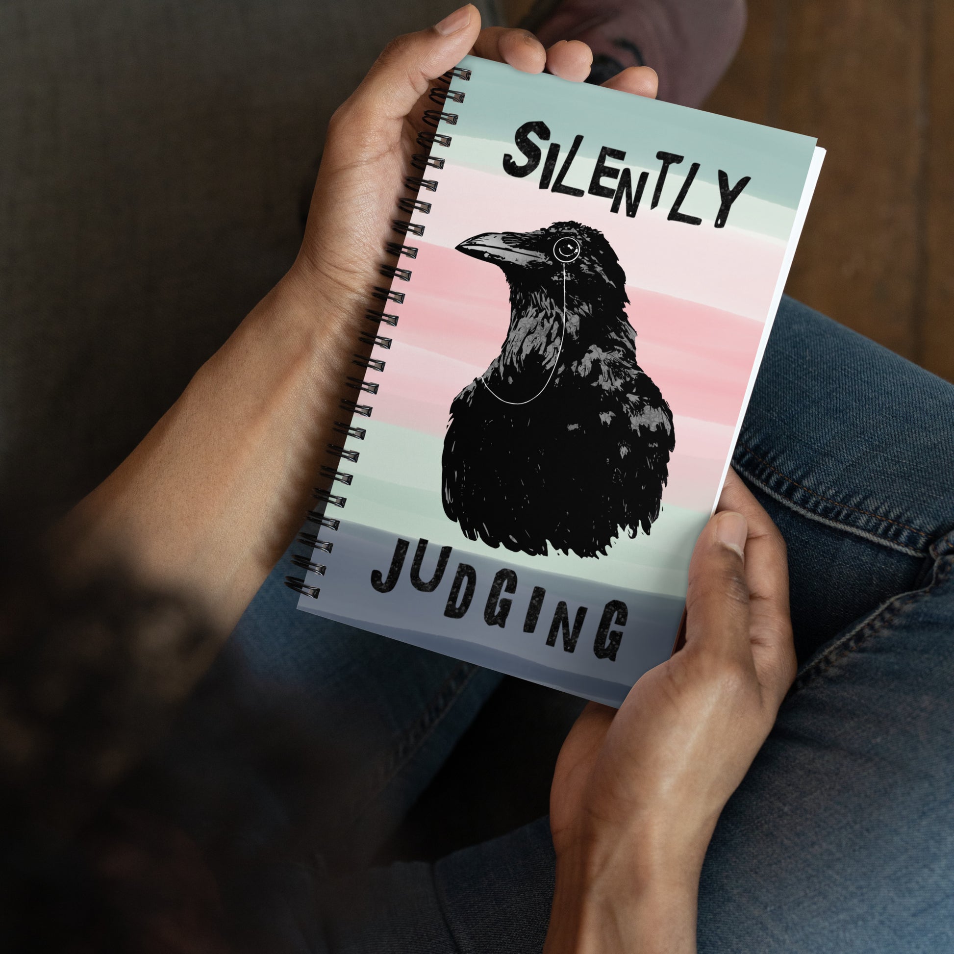 Spiral bound notebook with soft touch cover and 140 dotted pages. Cover features silently judging text and monocled crow on a striped background. Measures 5.5 inches by 8.5 inches. Shown in model's hands.