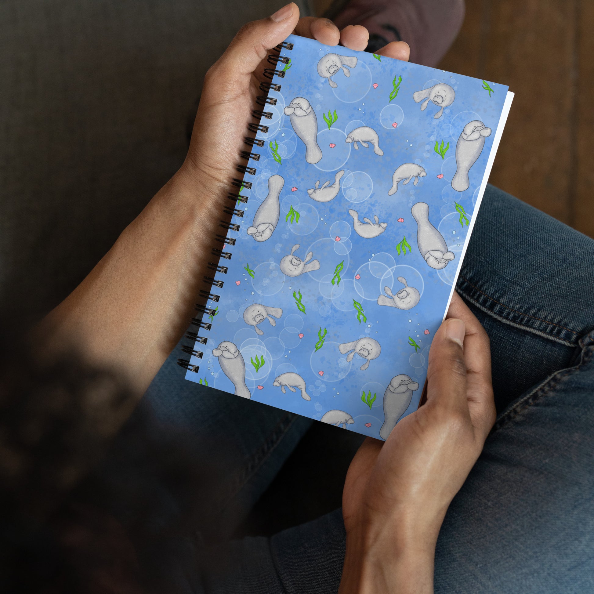Spiral bound notebook with soft touch cover and 140 dotted pages. Cover features cute illustrated manatees swimming in the water. Measures 5.5 inches by 8.5 inches. Shown in model's hands.