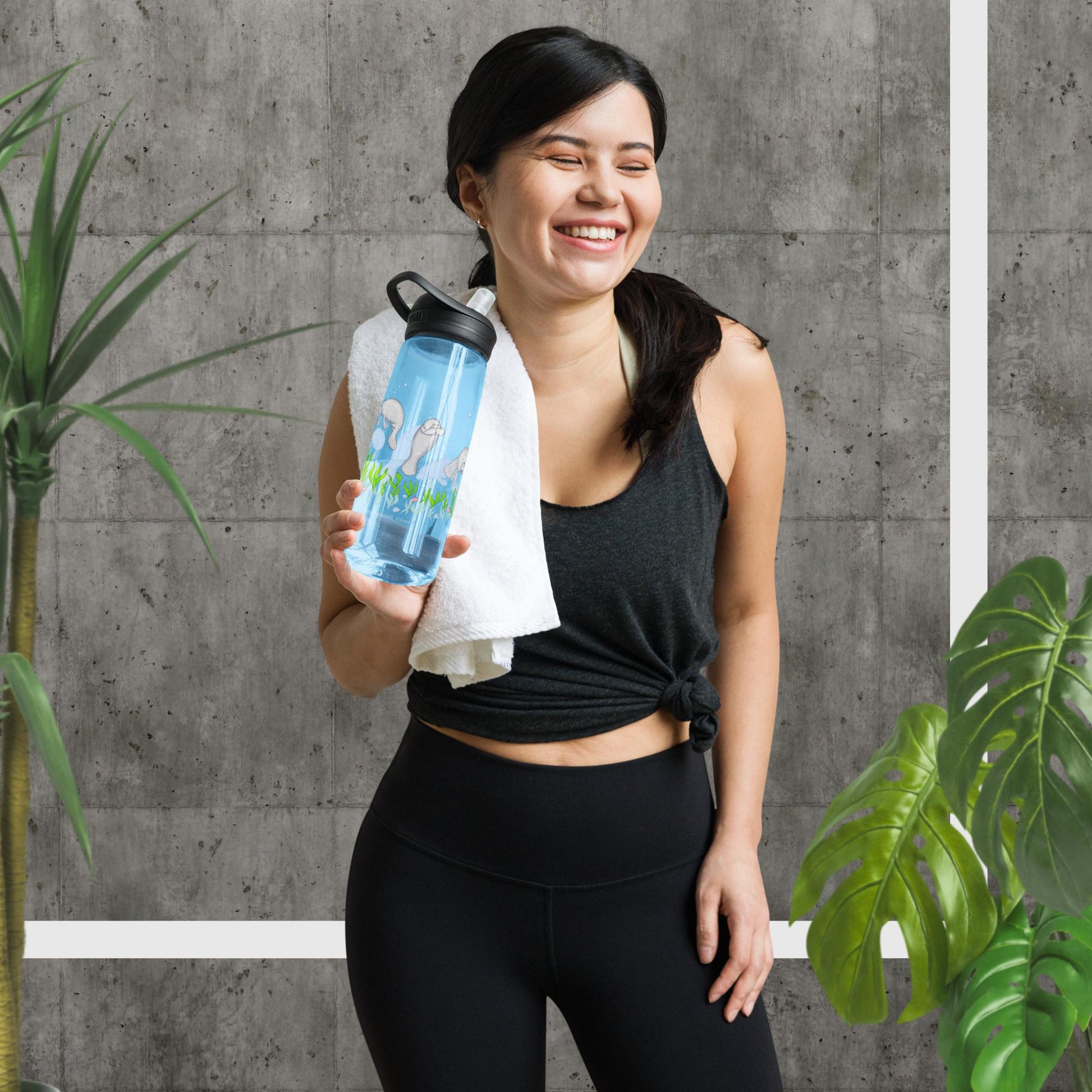25 ounce sports water bottle with spill proof lid and bite valve. Light blue stain and odor-resistant BPA-free plastic with manatee designs around the bottle, swimming above the seaweed and shells. Shown in gym model's hand by potted plants.