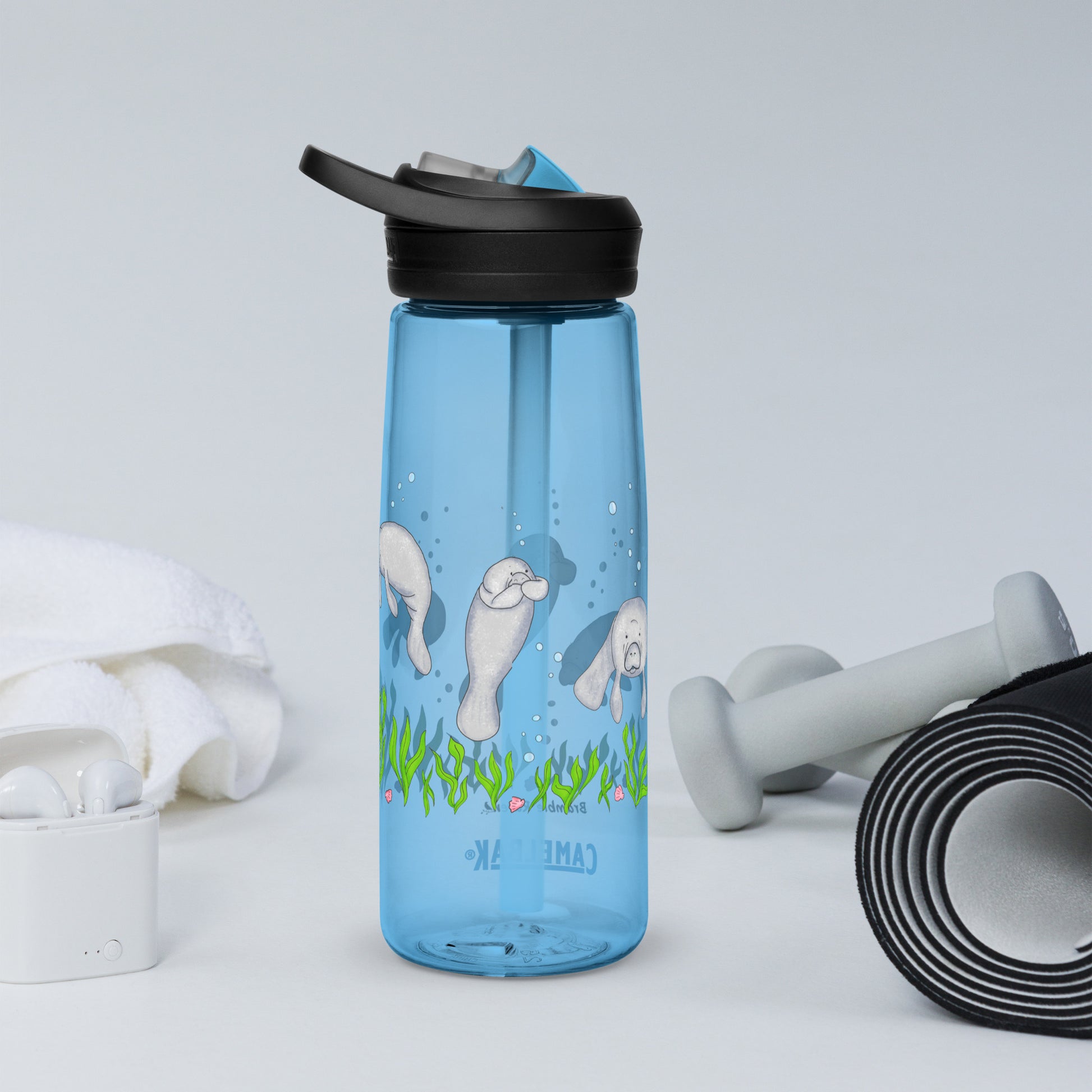 25 ounce sports water bottle with spill proof lid and bite valve. Ligh blue stain and odor-resistant BPA-free plastic with manatee designs around the bottle, swimming above the seaweed and shells. Shown on tabletop by yoga mat, weights, earbuds and towel.