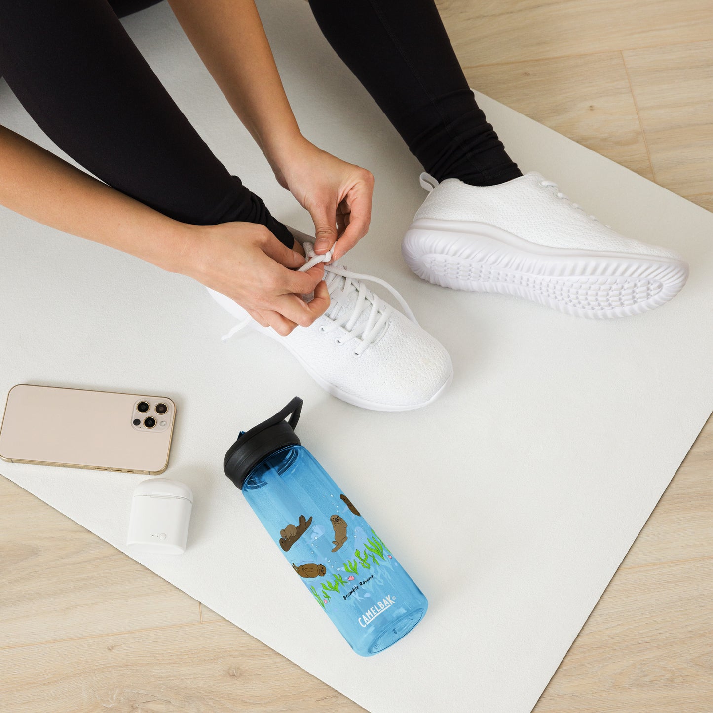 25 ounce sports water bottle with spill proof lid and bite valve. Light blue stain and odor-resistant BPA-free plastic with sea otter designs around the bottle, swimming above the seaweed and shells. Shown on yoga mat by model tying shoes.