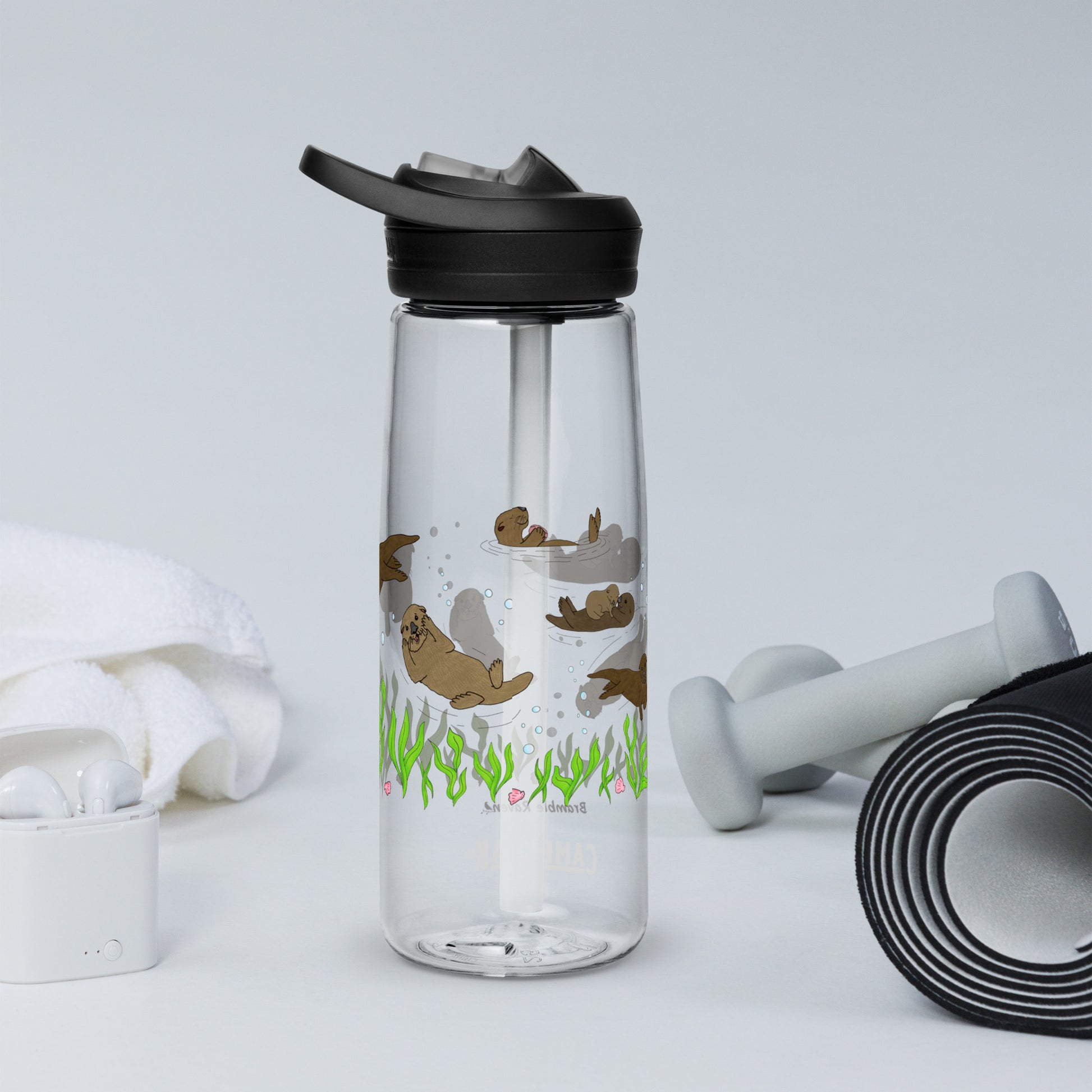 25 ounce sports water bottle with spill proof lid and bite valve. Clear stain and odor-resistant BPA-free plastic with sea otter designs around the bottle, swimming above the seaweed and shells. Shown on tabletop by yoga mat, towel, earbuds and weights.