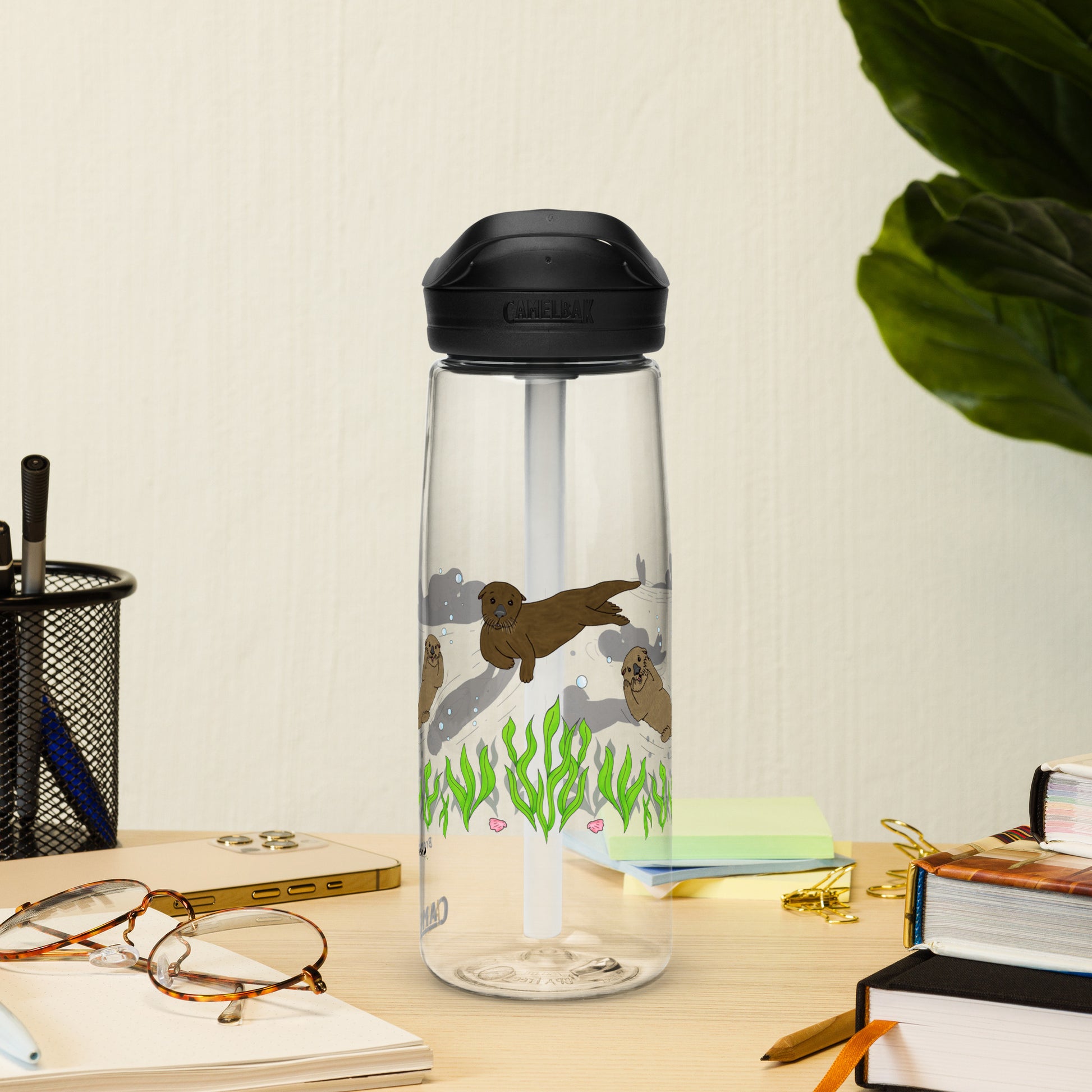 25 ounce sports water bottle with spill proof lid and bite valve. Clear stain and odor-resistant BPA-free plastic with sea otter designs around the bottle, swimming above the seaweed and shells. Shown on tabletop by books, pen holder, and glasses.