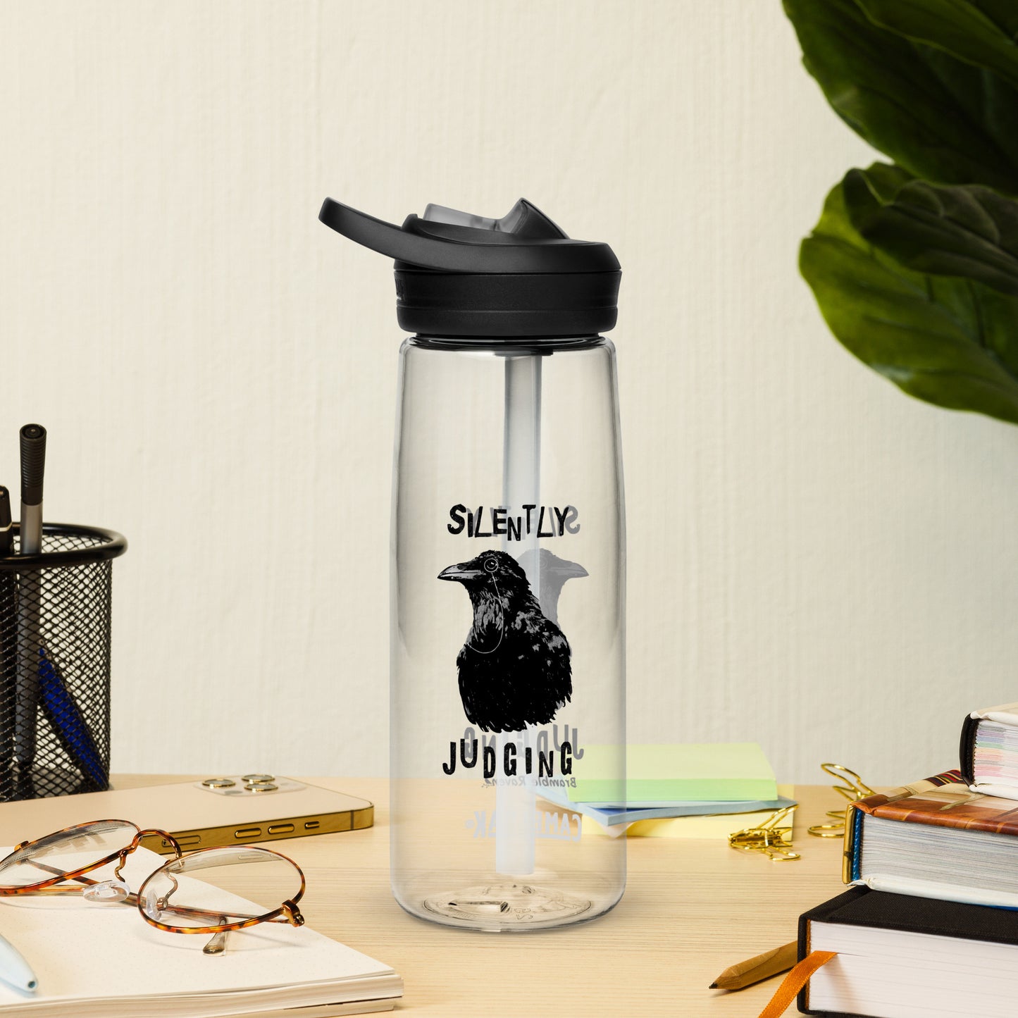 25 ounce sports water bottle with spill-proof lid and bite valve. Clear stain and odor-resistant BPA-free plastic. Features double-sided design of Silently Judging Crow with his monocle. Shown on desk by books, pen holder, and glasses.