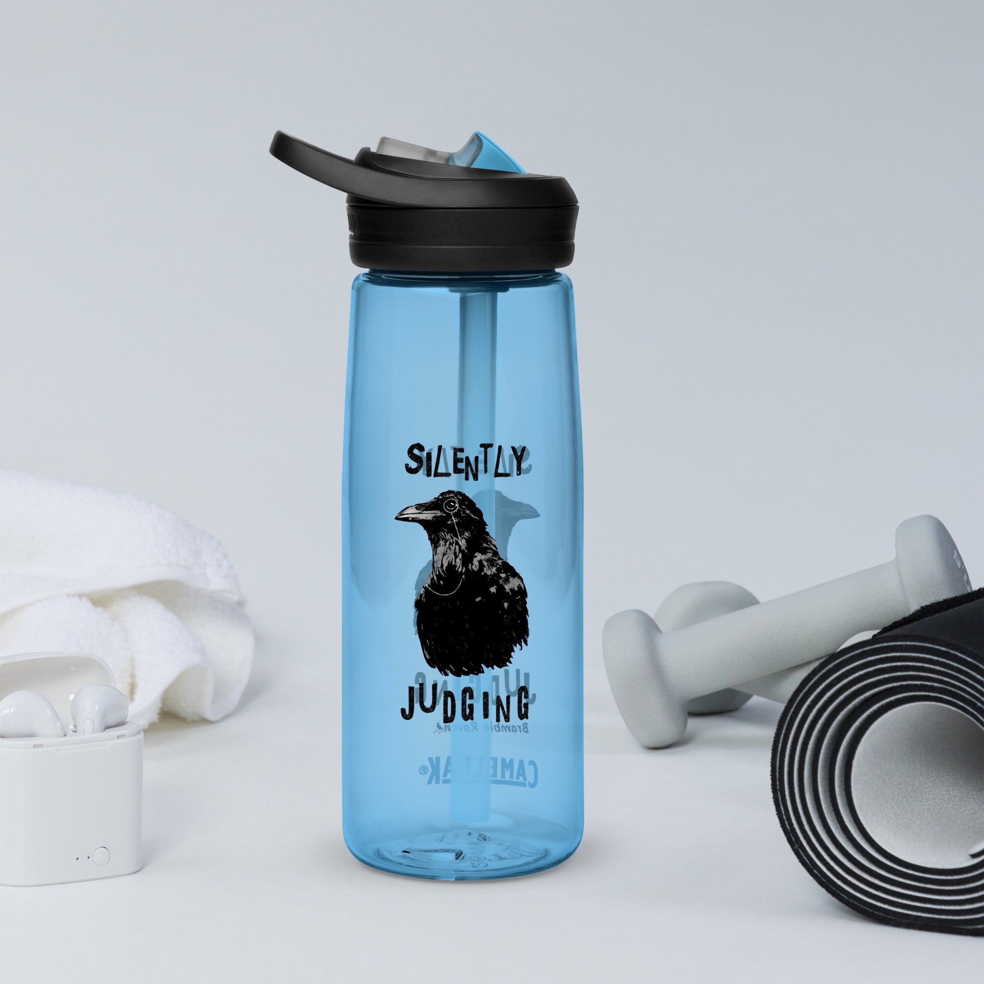25 ounce sports water bottle with spill-proof lid and bite valve. Light blue stain and odor-resistant BPA-free plastic. Features double-sided design of Silently Judging Crow with his monocle. Shown by yoga mat, weights, and towel.