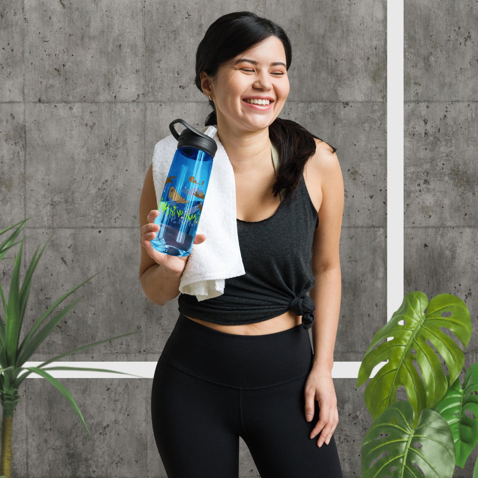 25 ounce sports water bottle with spill proof lid and bite valve. Dark blue stain and odor-resistant BPA-free plastic with sea otter designs around the bottle, swimming above the seaweed and shells. Shown in smiling gym model's hands next to house plants.