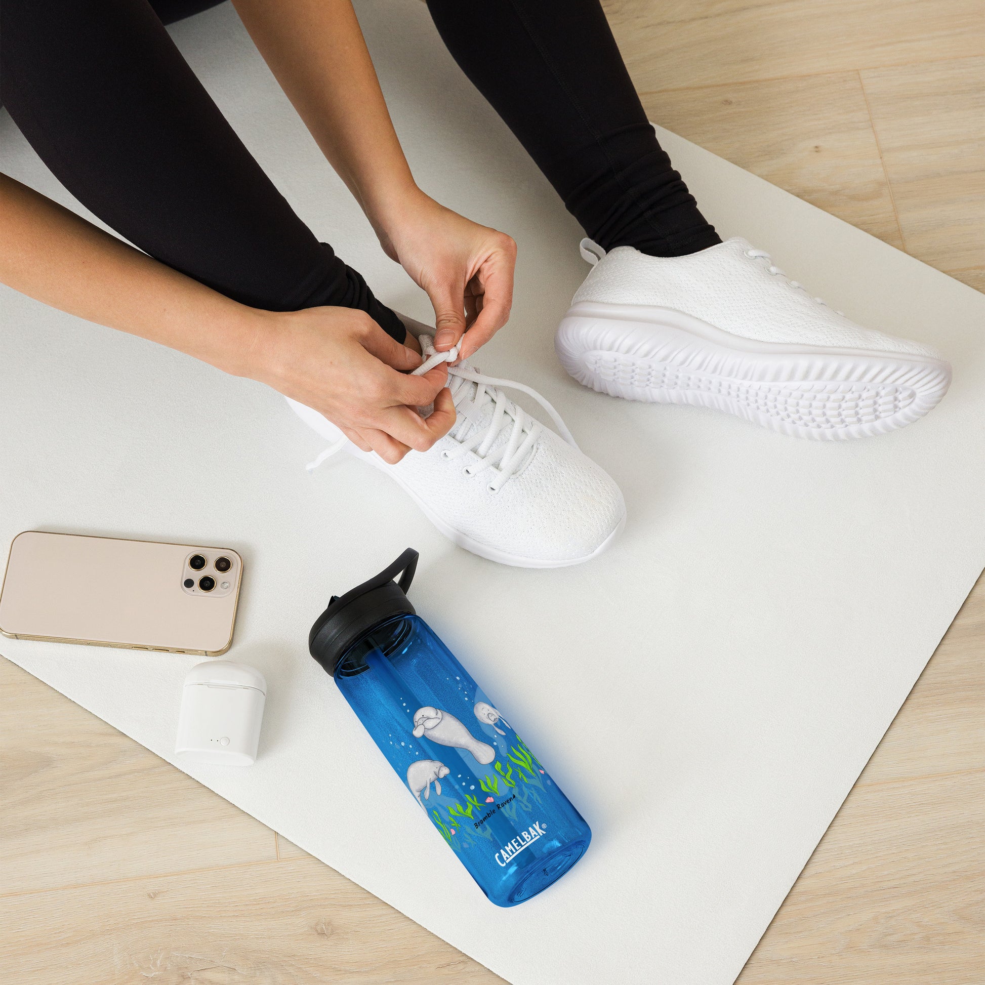 25 ounce sports water bottle with spill proof lid and bite valve. Dark blue stain and odor-resistant BPA-free plastic with manatee designs around the bottle, swimming above the seaweed and shells. Shown on yoga mat by model tying shoes.