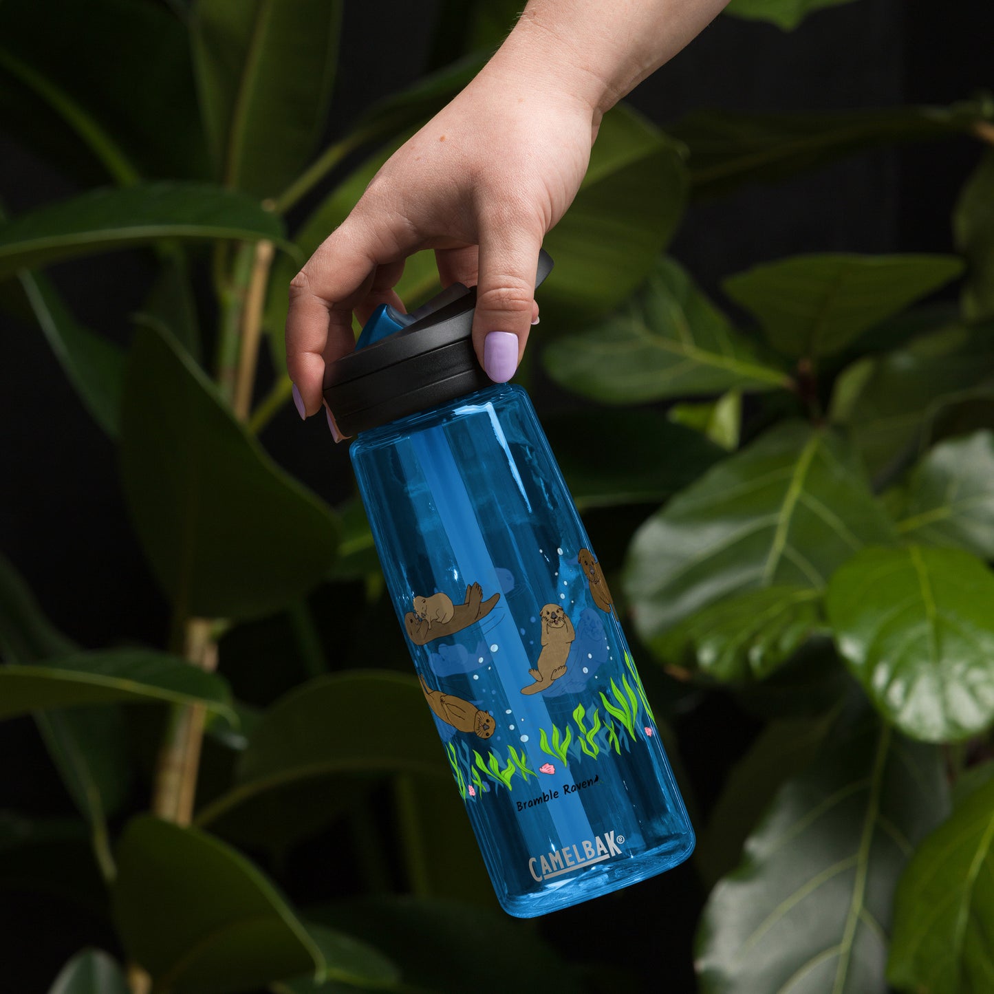 25 ounce sports water bottle with spill proof lid and bite valve. Dark blue stain and odor-resistant BPA-free plastic with sea otter designs around the bottle, swimming above the seaweed and shells. Shown in model's hand by plants.