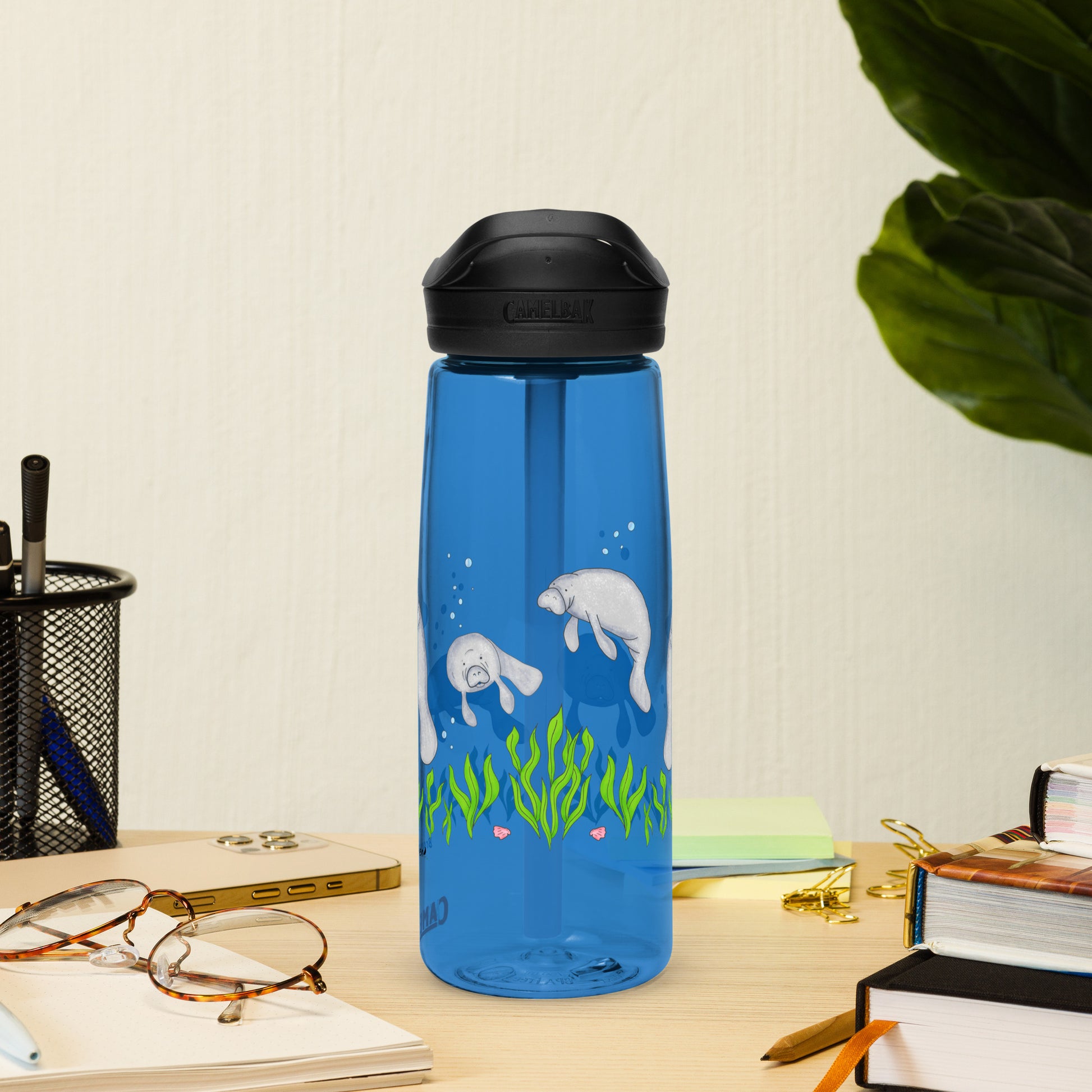 25 ounce sports water bottle with spill proof lid and bite valve. Dark blue stain and odor-resistant BPA-free plastic with manatee designs around the bottle, swimming above the seaweed and shells. Shown on tabletop by books, pen holder and glasses.