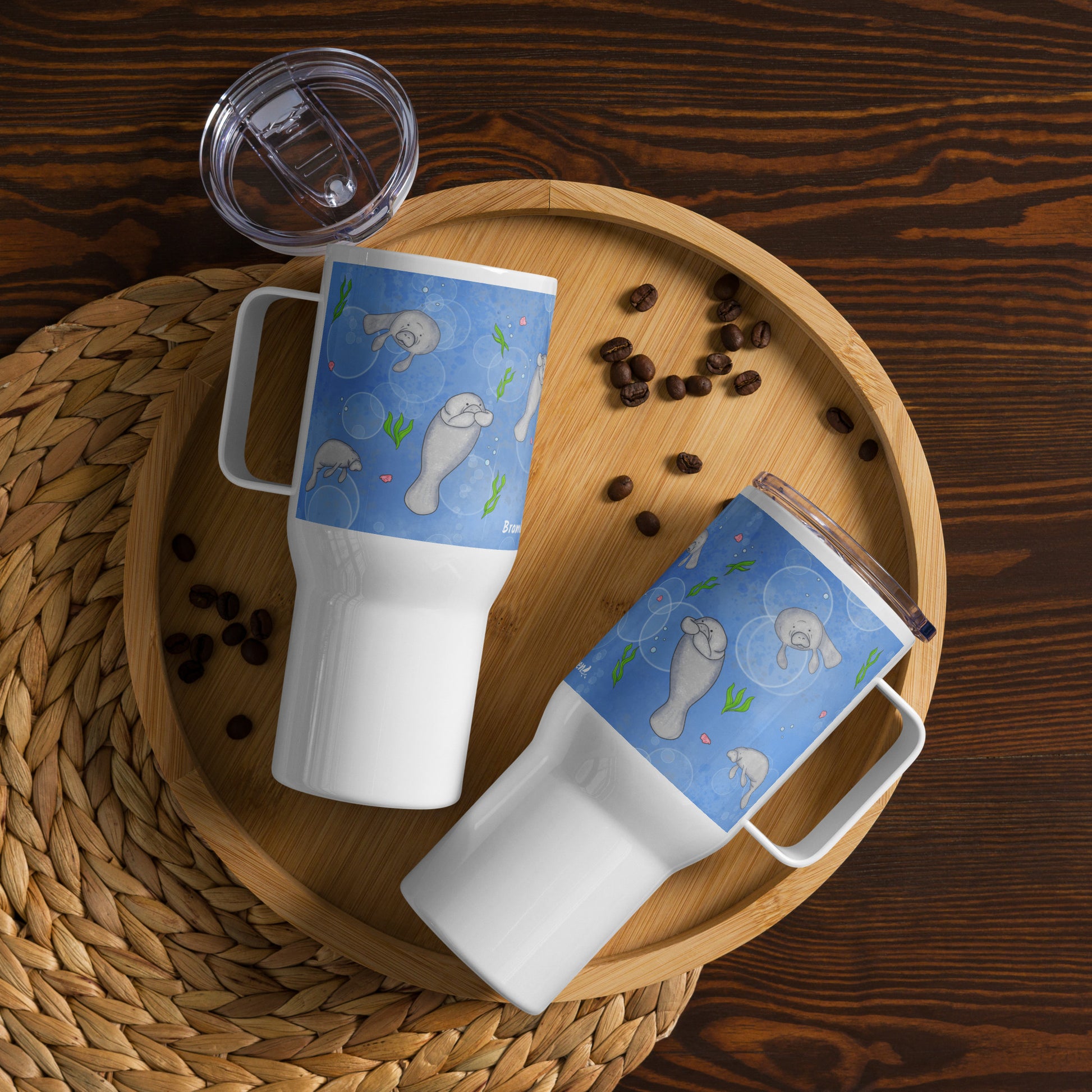 Stainless steel travel mug with handle. Holds 25 ounces of hot or cold drinks. Has wraparound illustrated manatees design. Fits most car cup holders. Comes with spill-proof BPA-free plastic lid. Image shows two mugs on wooden serving tray by coffee beans.