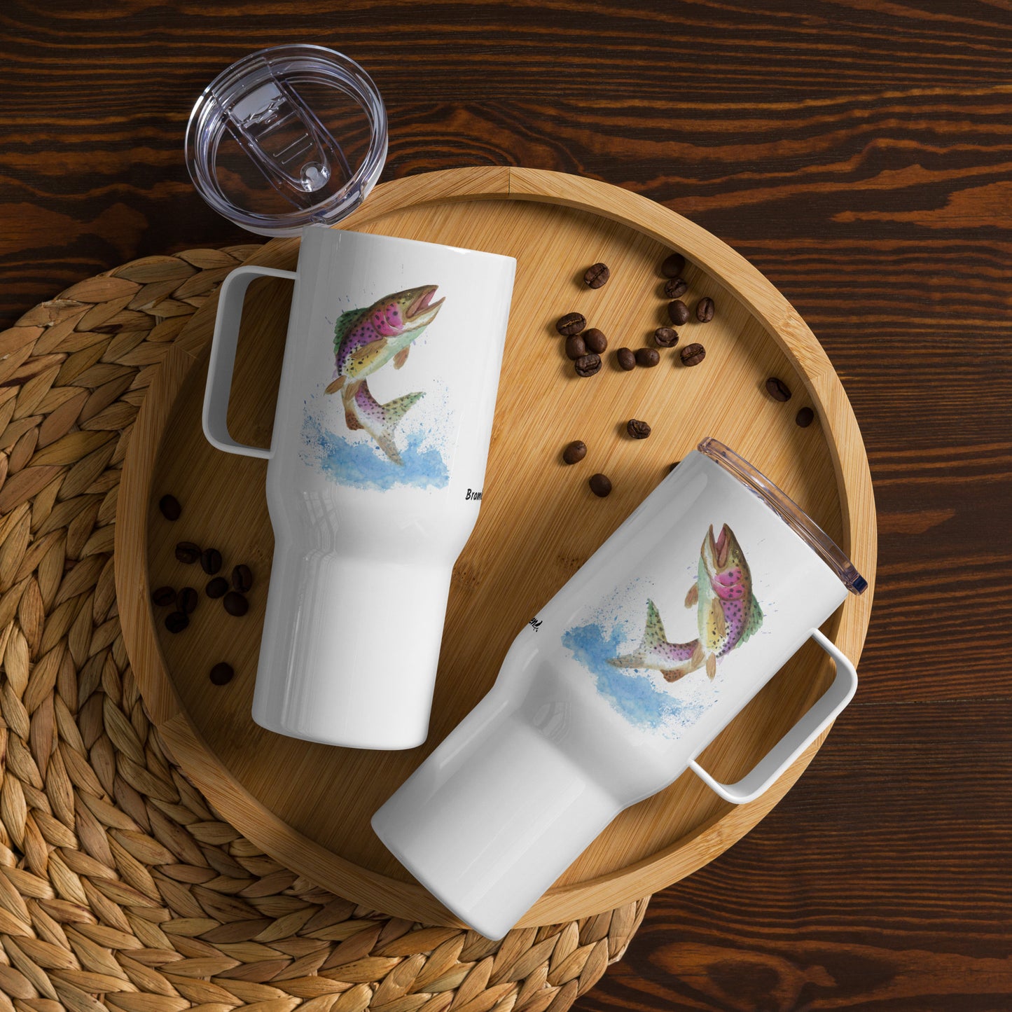 Stainless steel travel mug with handle. Holds 25 ounces of hot or cold drinks. Has print of watercolor rainbow trout fish on both sides. Fits most car cup holders. Comes with spill-proof BPA-free plastic lid. Image shows two mugs on wooden tray by coffee beans.