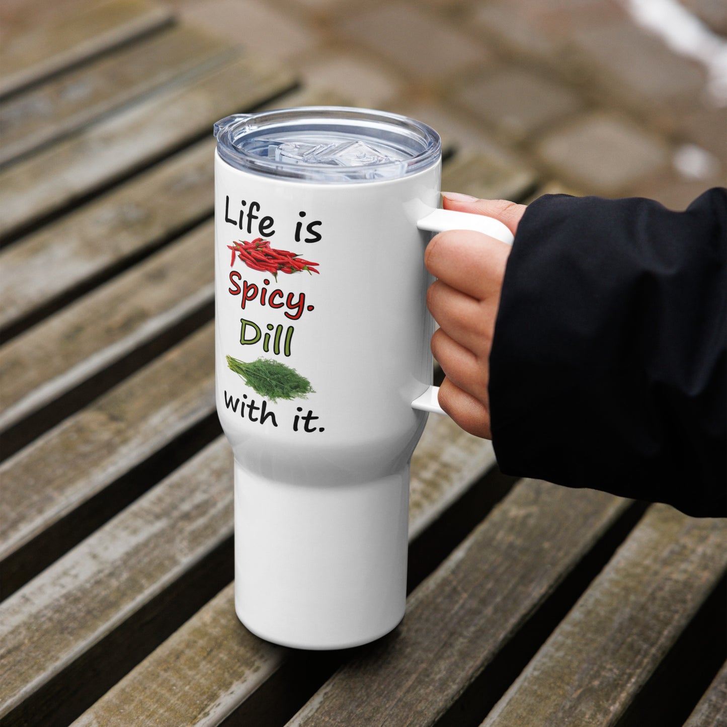 Stainless steel travel mug with handle. Holds 25 ounces of hot or cold liquids. Fits most car cup holders. Features double-sided phrase: Life is spicy. Dill with it, and accompanying chili pepper and dill weed images. Comes with spill proof plastic lid. Shown in model's hand on wooden table.